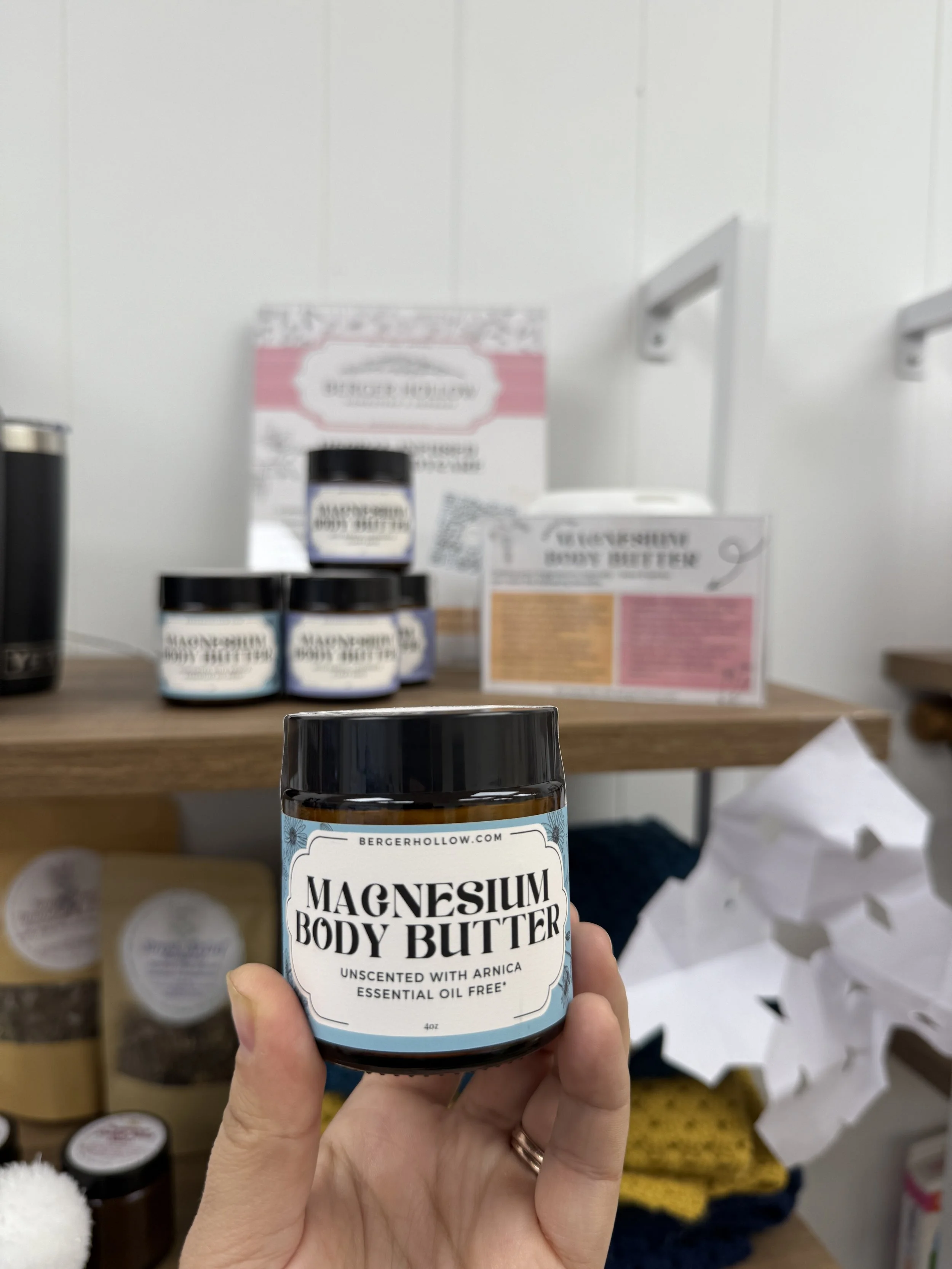 A hand holding a jar of magnesium body butter in a store, with other jars and product displays blurred in the background.