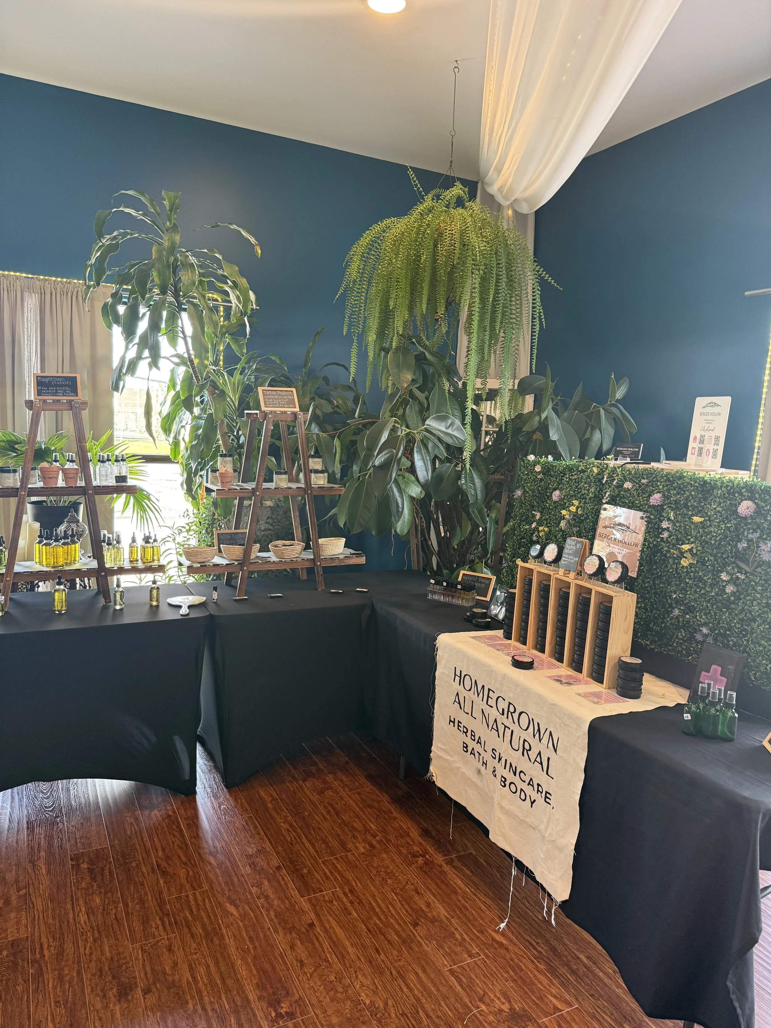 Display of herbal products and skincare items at a booth with a beachy sign saying 'Homegrown All Natural Herbal Skincare, Bath & Body' in an indoor setting with plants and black tablecloths.