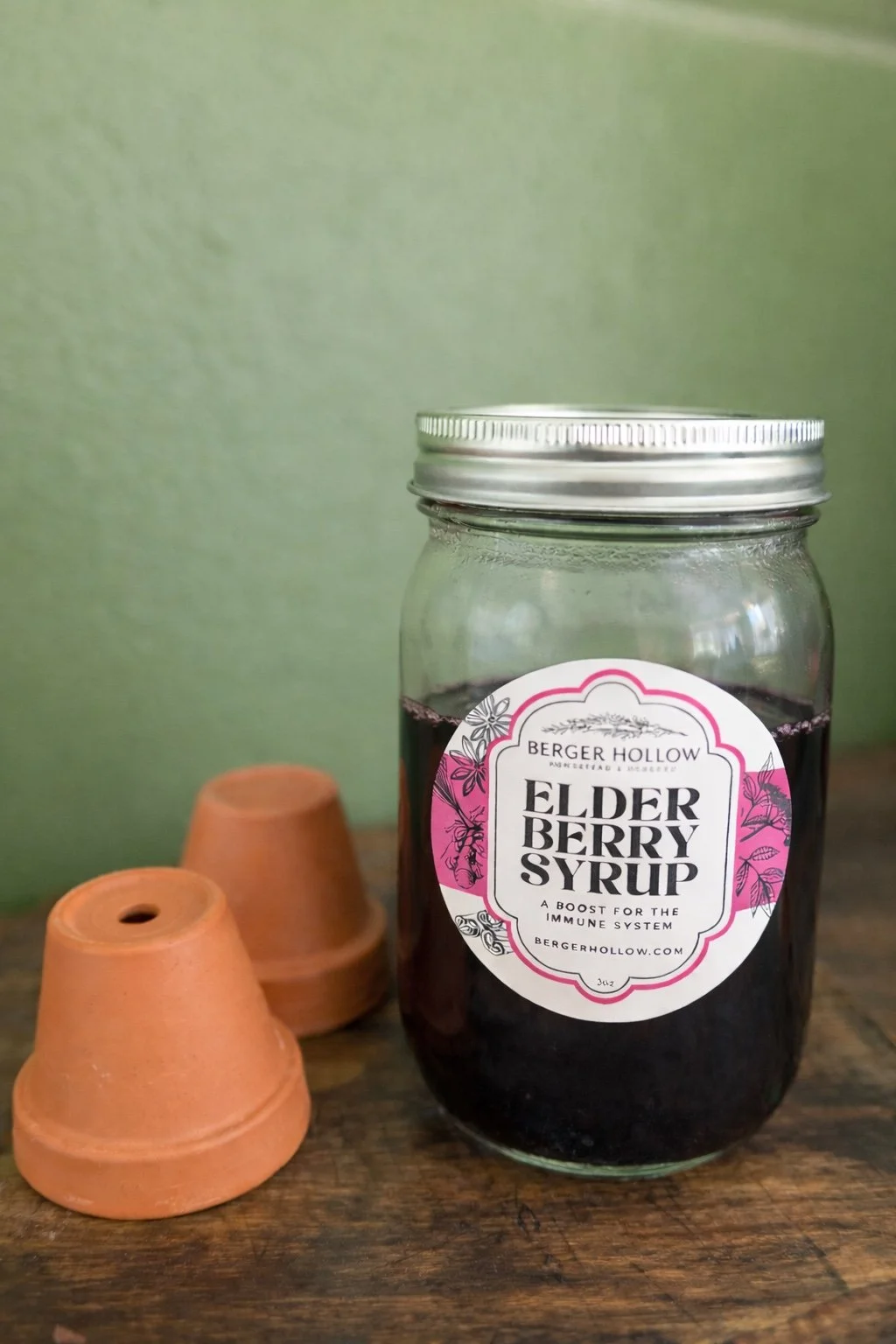 A glass jar of elderberry syrup with a metal lid, placed on a wooden surface, next to two small terracotta pots, against a green wall background.