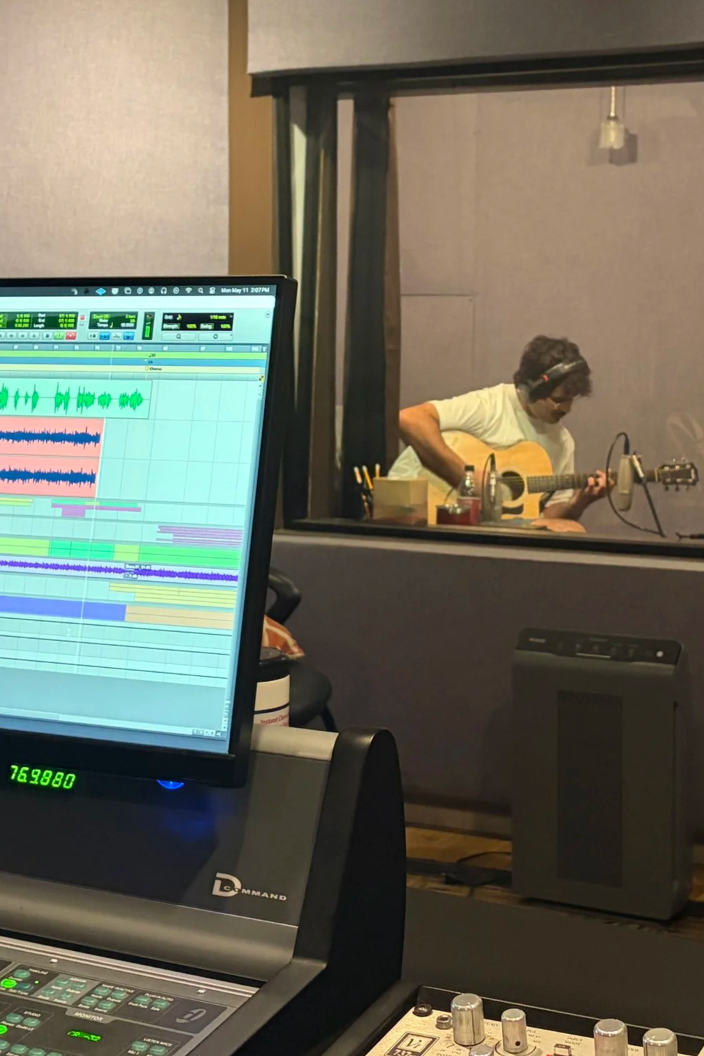 A person wearing headphones playing an acoustic guitar in a recording studio, observed through a glass window from a control room with audio equipment and a computer screen showing a digital audio workstation.