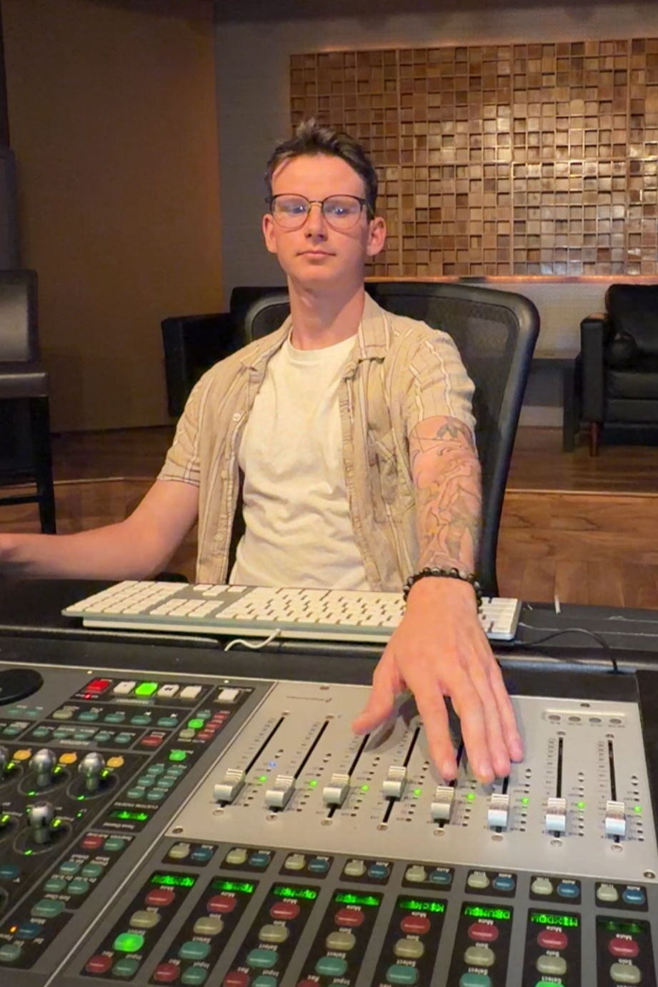 A young man with glasses and tattoos sitting at a music studio mixing console.