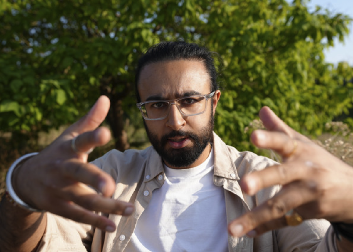 Man with glasses and a beard looking at the camera outdoors with green trees in the background.