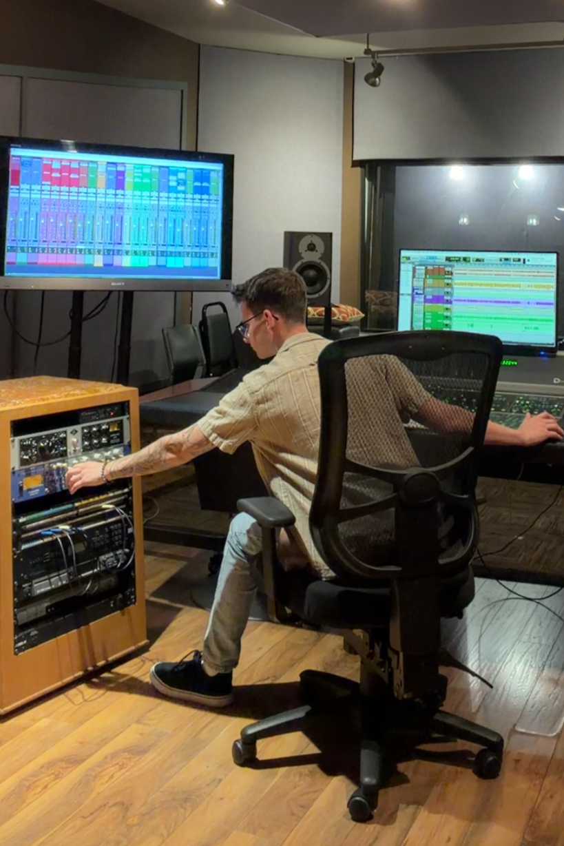 Two men working in a music studio with mixing equipment, large monitors displaying digital audio workstations, and acoustic panels on the walls.