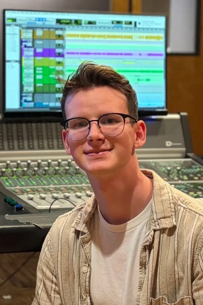 A young man wearing glasses and a striped beige shirt sitting in front of a professional audio mixing console and a computer screen showing digital audio workstation software.