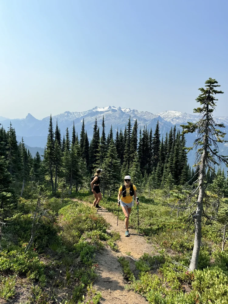 Two trail runners climbing a subalpine trail with snowy Cascade peaks behind them