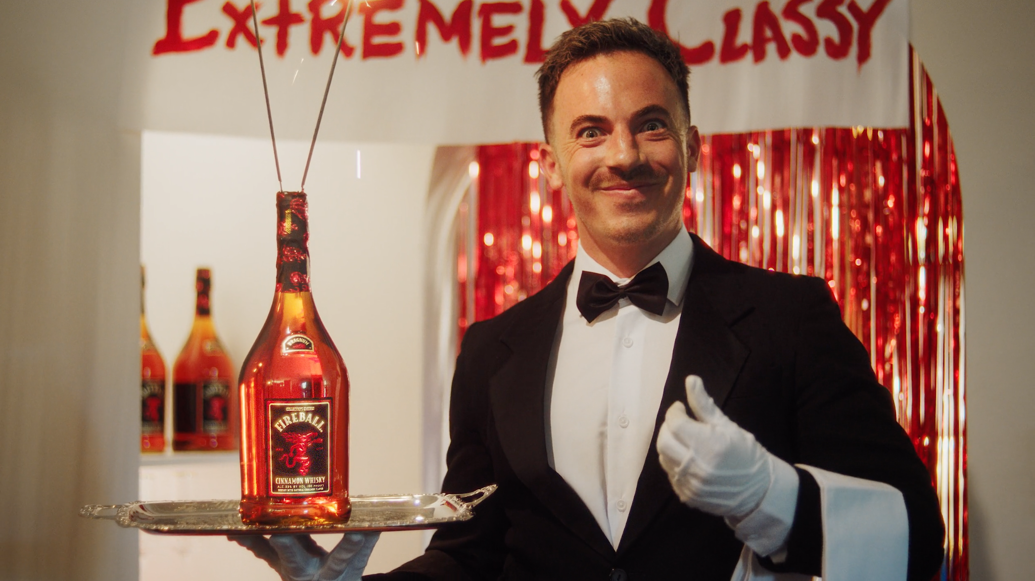 Man dressed as a tuxedo butler serving bottles of Fireball cinnamon whisky at an event with a red and white 'Extremely Classy' banner in the background.