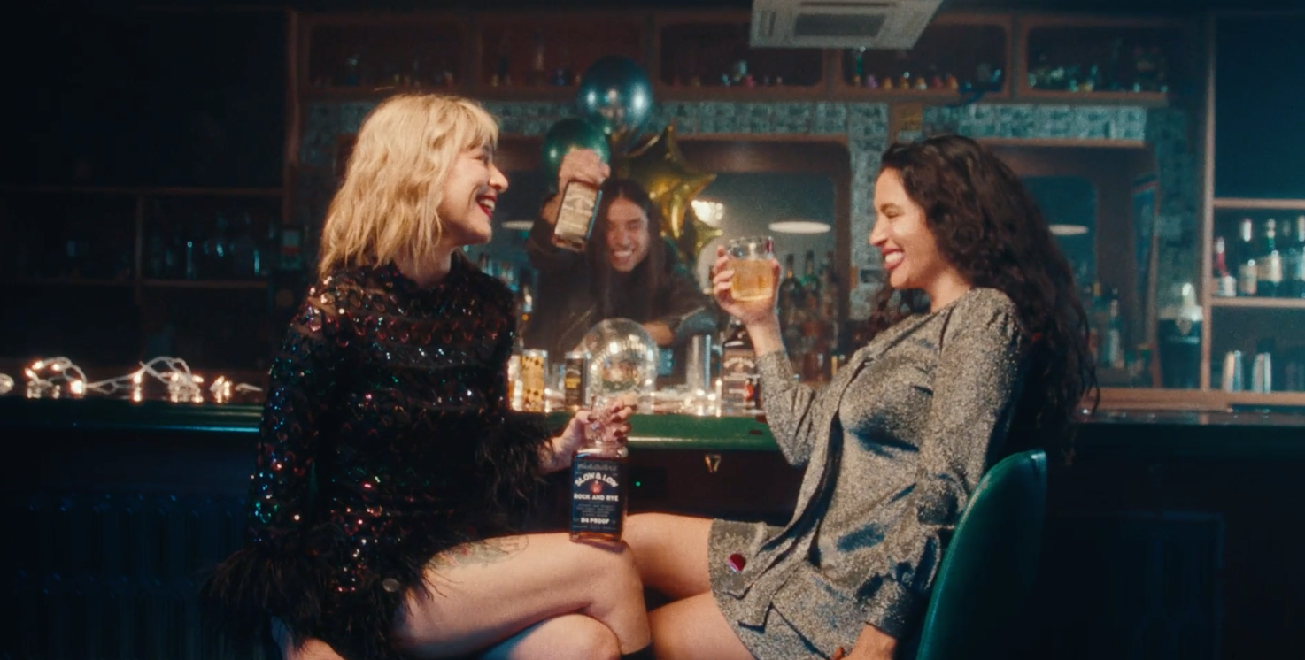 Two women sitting at a bar, laughing and toasting with drinks, with a bartender serving behind them, decorated with balloons and lights for a celebration.
