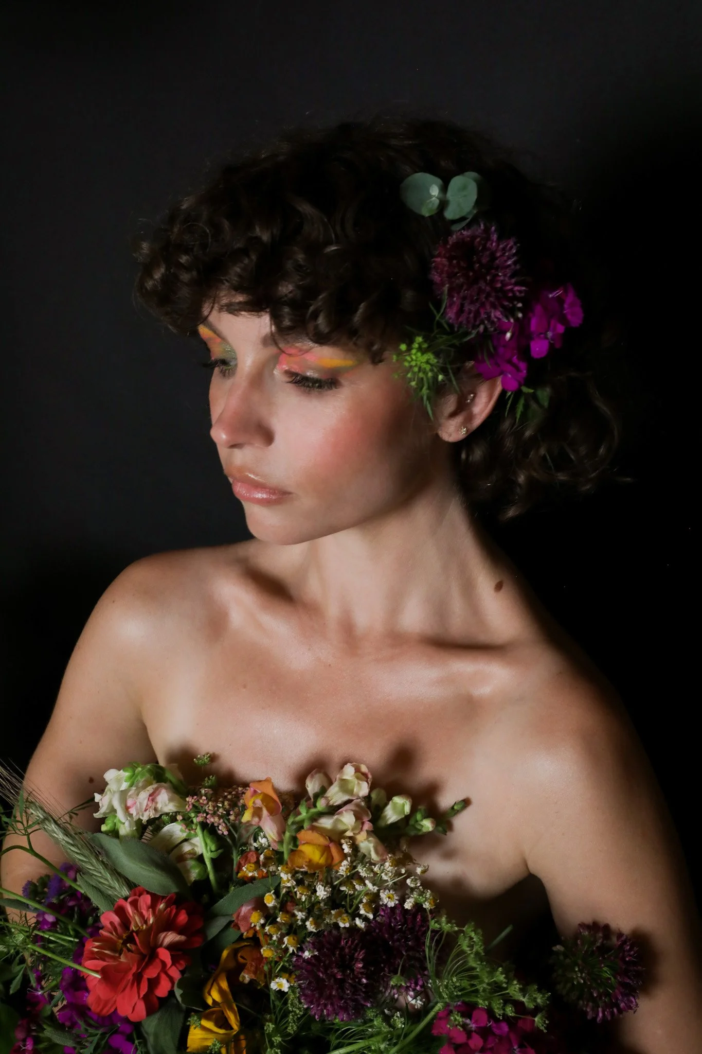 A woman with curly hair adorned with purple and green flowers, holding a large bouquet of various colorful flowers including pink, red, yellow, and white blossoms against a black background.
