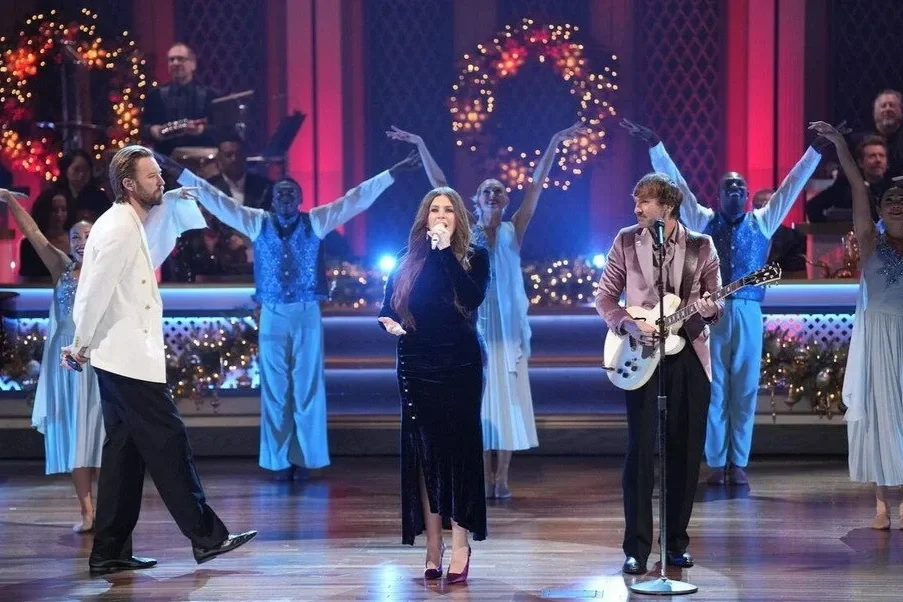 Performers singing and playing guitar on stage decorated with Christmas wreaths and fairy lights, with dancers in festive attire in the background.