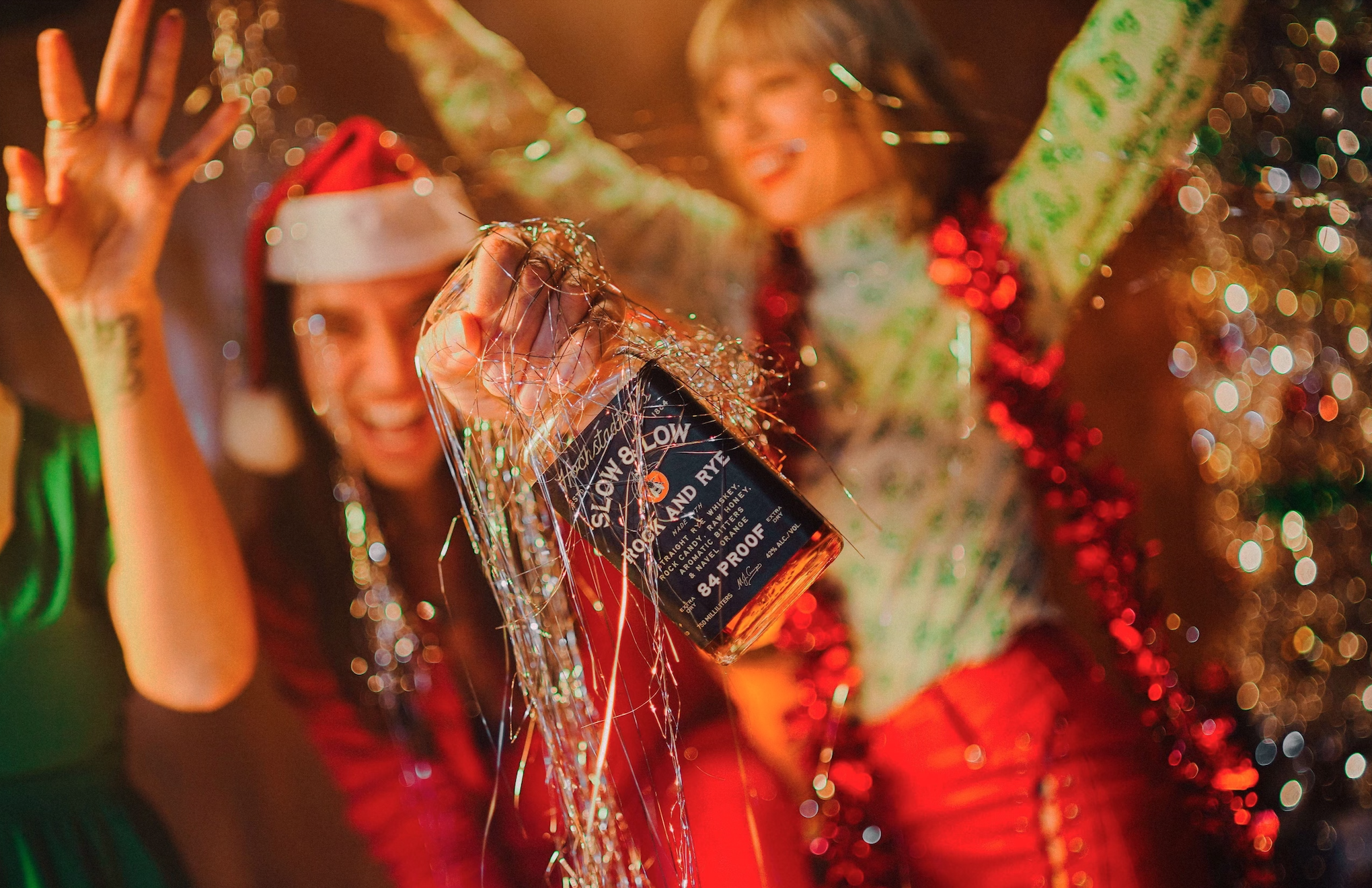 Person holding a bottle of whiskey, with people celebrating in the background, colorful lights, and festive decorations, including tinsel and wrapped presents.