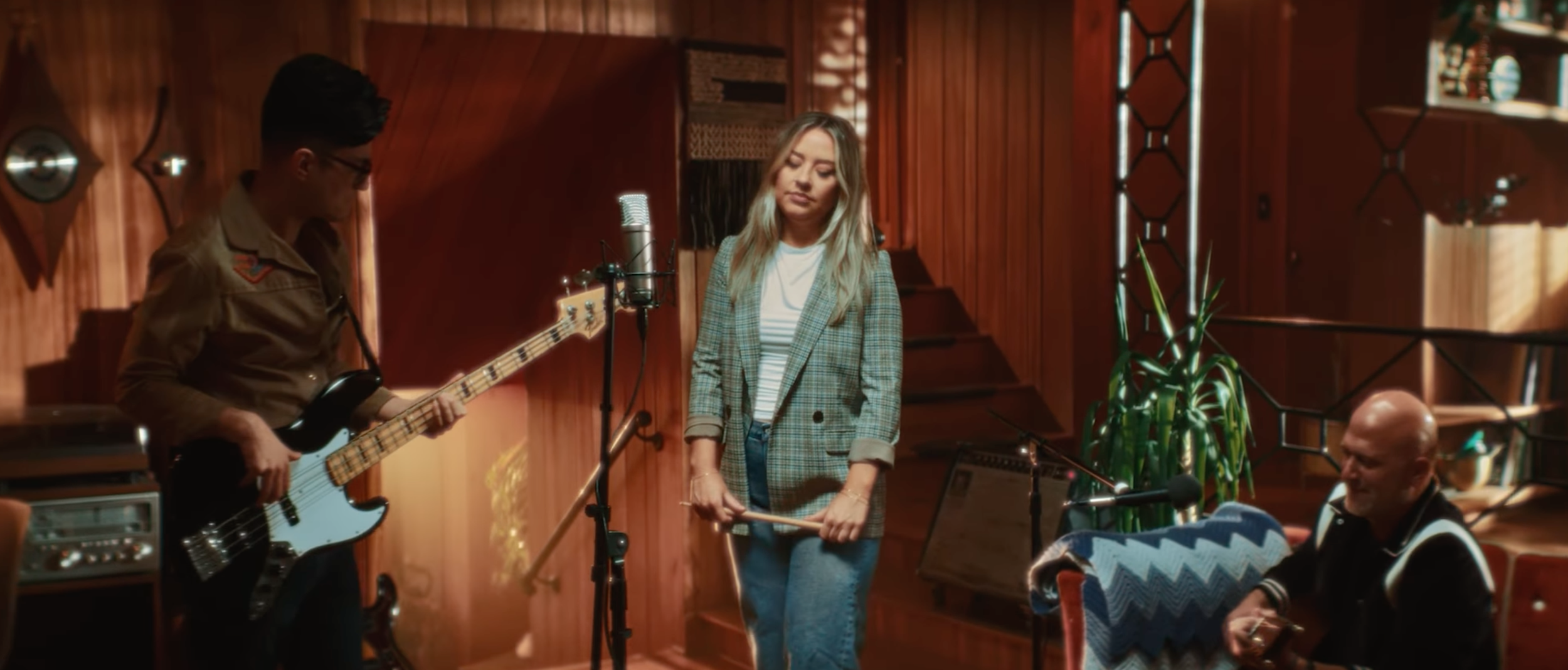 Three musicians practicing in a wooden-paneled studio; a young man playing bass, a woman holding drumsticks, and an older man seated with a guitar.
