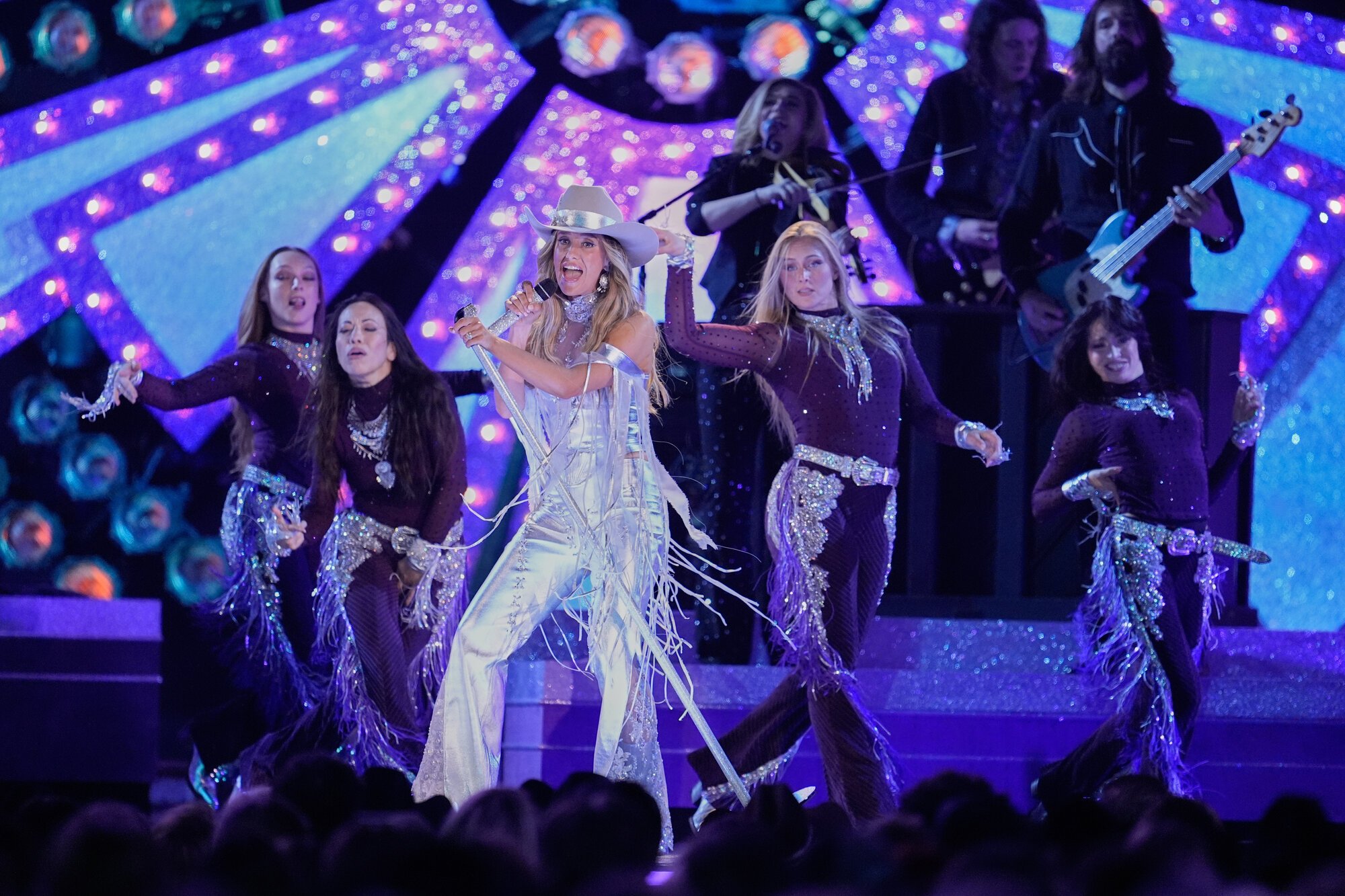 A stage performance featuring a woman in white cowboy attire singing into a microphone, surrounded by backup dancers in purple costumes and backup musicians playing guitar against a colorful, illuminated backdrop.