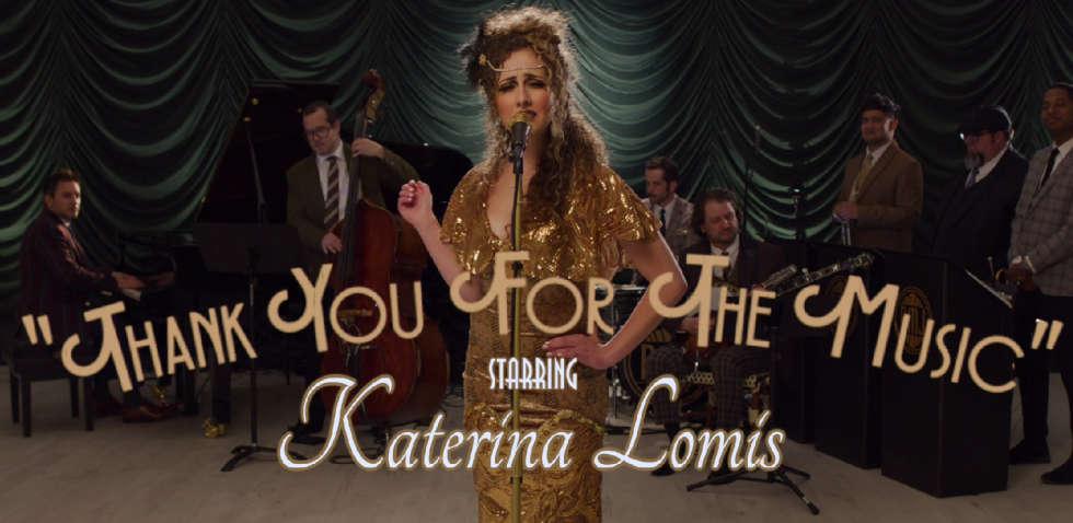 A woman with curly hair and a gold dress singing into a microphone, with a jazz band playing behind her.