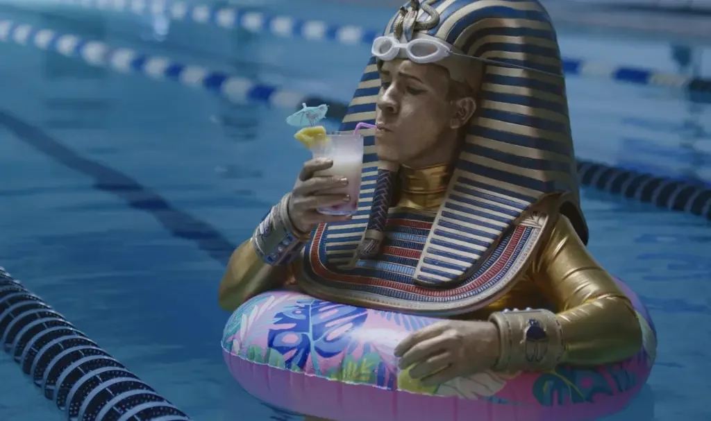 A person dressed as an ancient Egyptian pharaoh, wearing a striped headdress and gold costume, sitting in a swimming pool with a colorful inflatable ring, drinking a cocktail with a small umbrella and decorative straw.