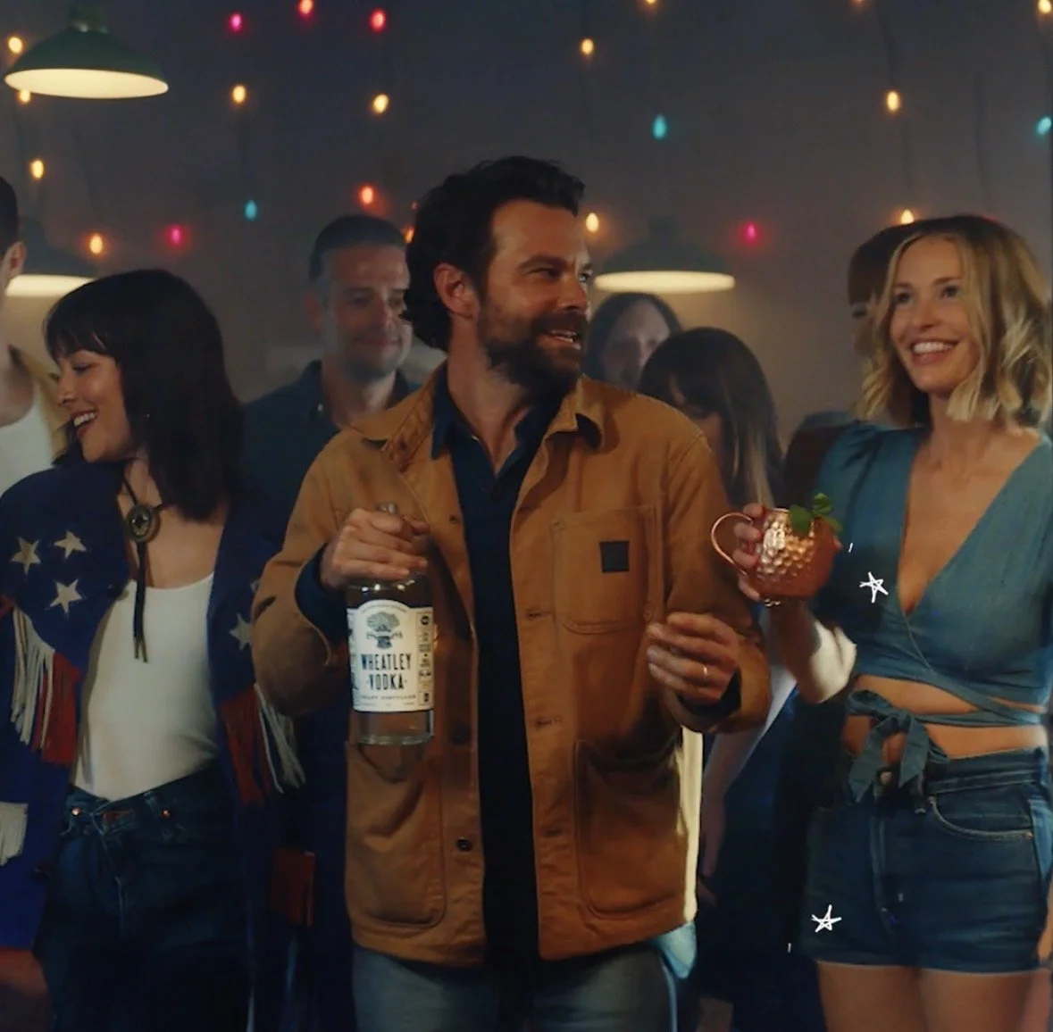 People enjoying a lively party, with colorful string lights and a festive atmosphere, some holding drinks and smiling.