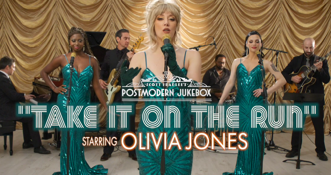 Olivia Jones in a teal sequin dress singing into a microphone on stage with a band and backup singers, gold curtain backdrop.