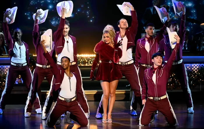 A woman in a red dress and high heels performing a dance on stage with eight male dancers in maroon track suits and white hats.