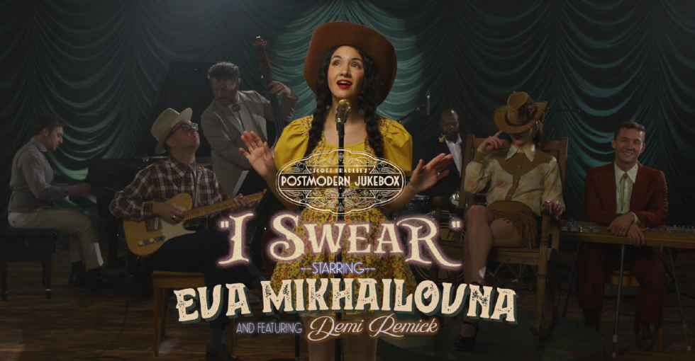 A musical performance on stage with a group of musicians and singers, woman in yellow dress and wide-brimmed hat at microphone, backdrop with teal curtains, overlay text for a jukebox song titled 'I Swear' by Eva Mikhailovna featuring Demi Demick.