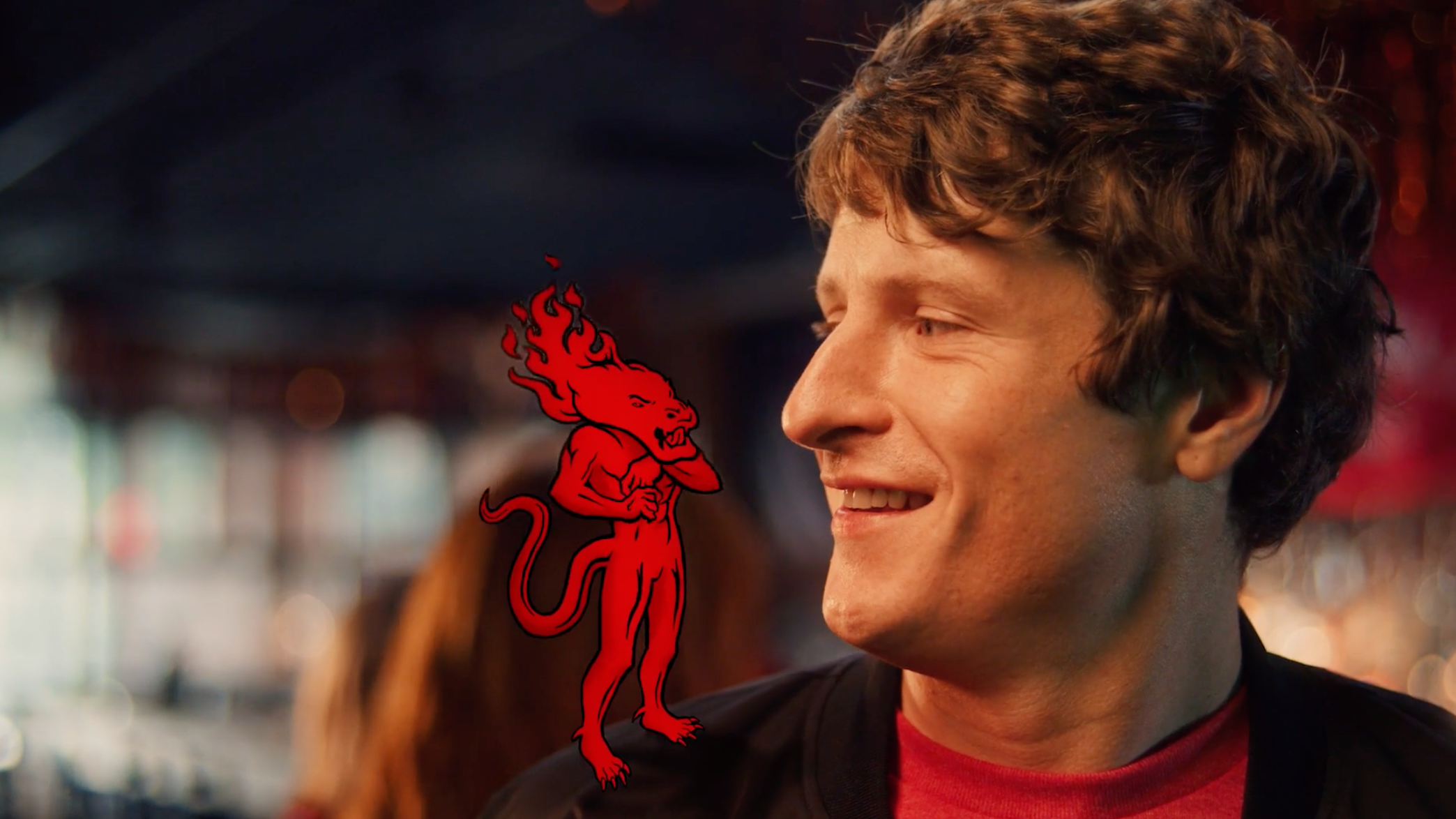 Close-up of a smiling young man with curly brown hair looking to the right, wearing a red shirt and dark jacket, with a red devil graphic overlay on his shoulder in a blurred background.