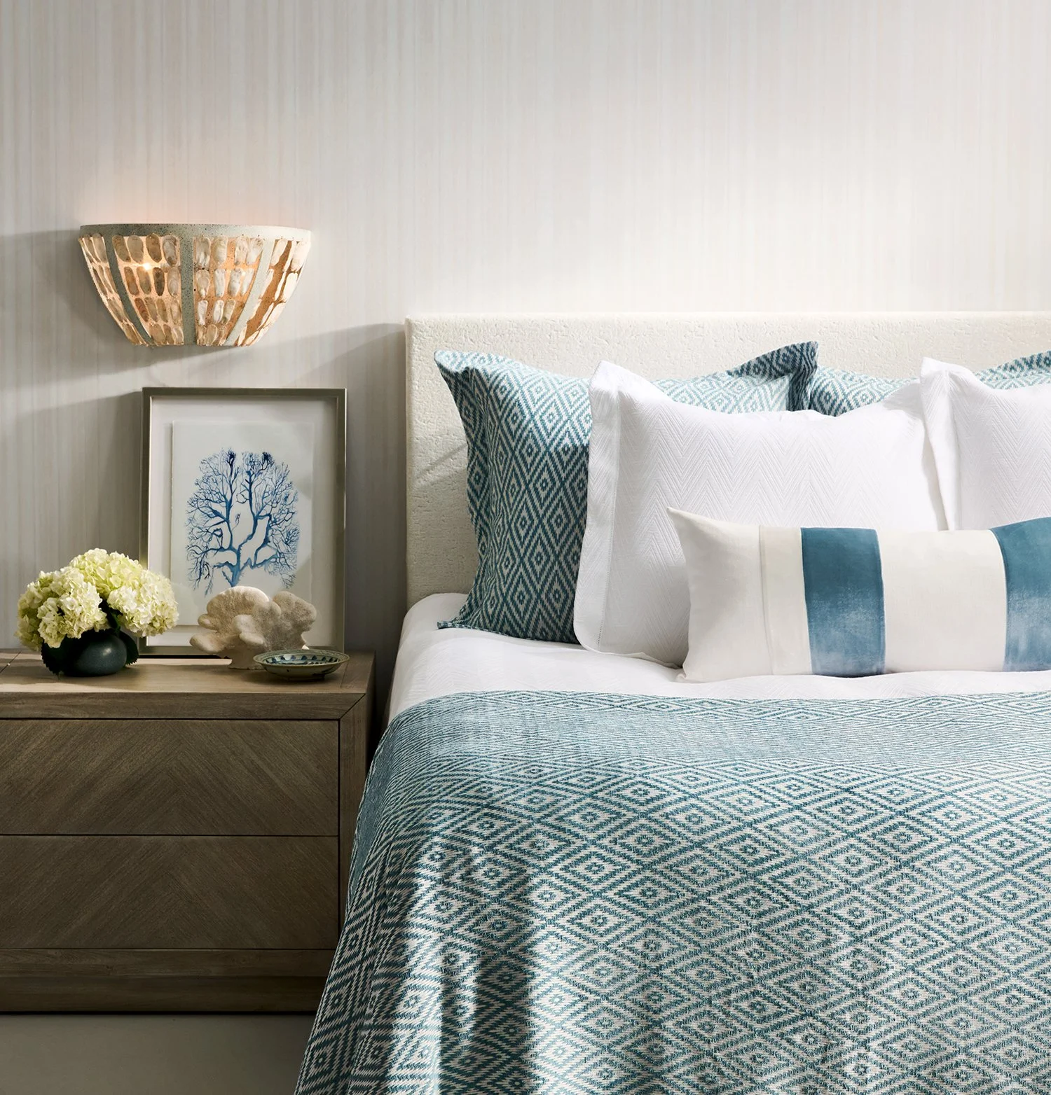 Bedroom with white and blue decor, including pillows and bedspread, a wooden nightstand with flowers, art, and a wall sconce.