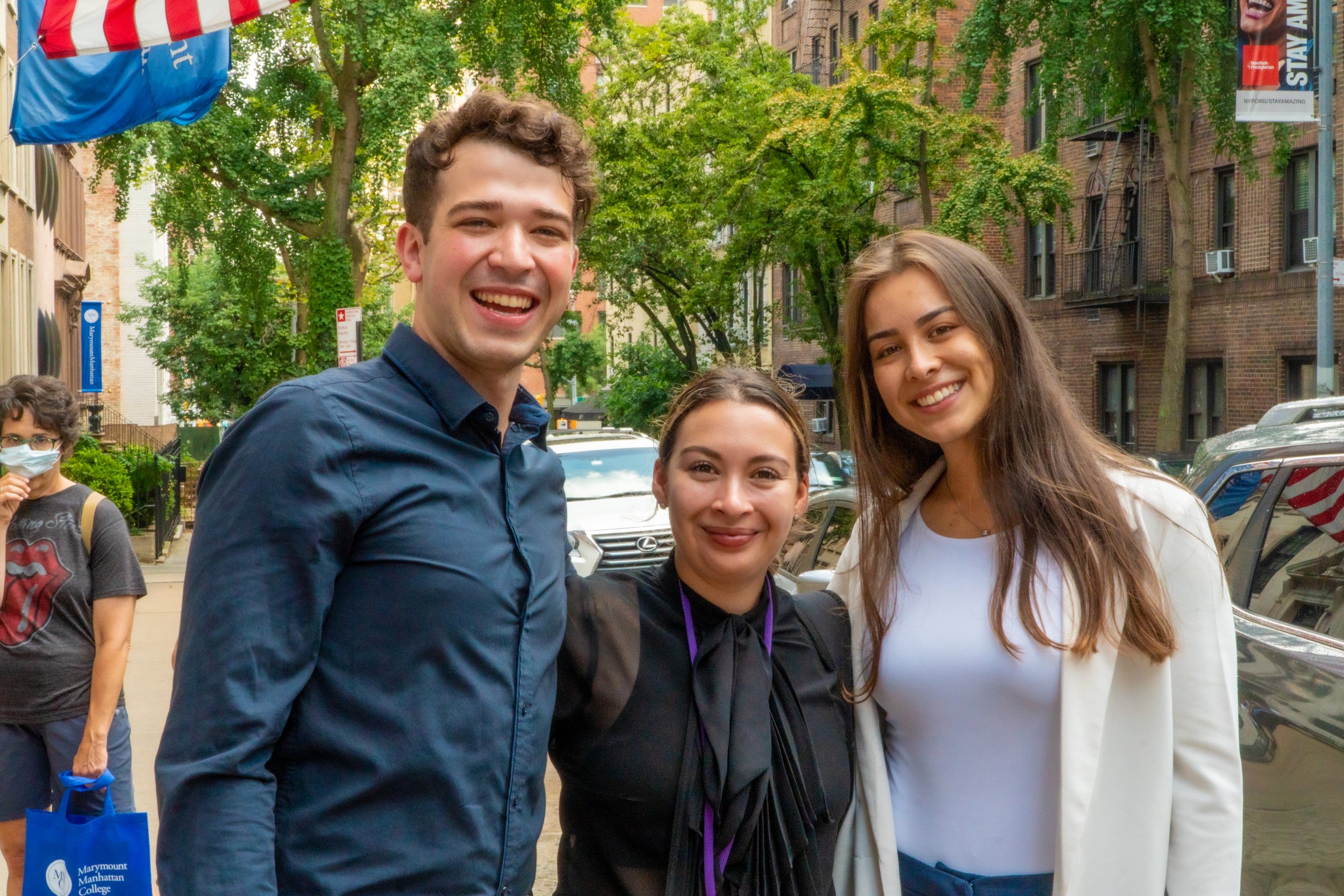 Three people smiling and posing for a photo on a city sidewalk with trees and buildings in the background.