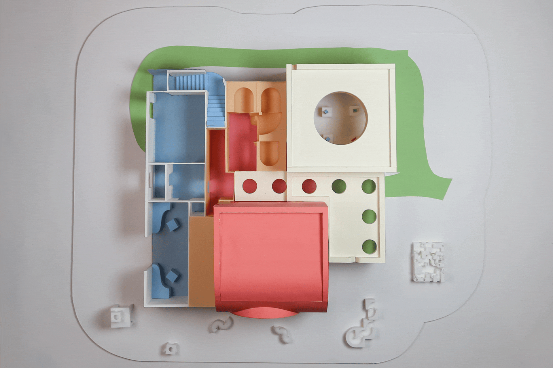 Top-down view of a colorful dollhouse with multiple rooms, including blue, red, beige, and white sections, placed on a white surface with small white accessories nearby.