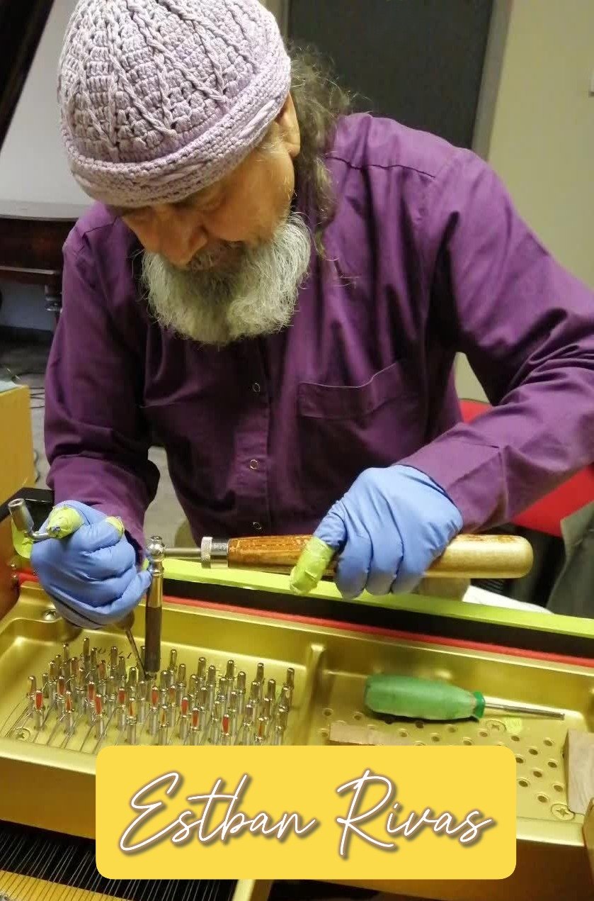 An elderly man with a white beard wearing a pink knit cap, purple shirt, and blue gloves works on a piano string hammer using a small tool, seated at a workbench.