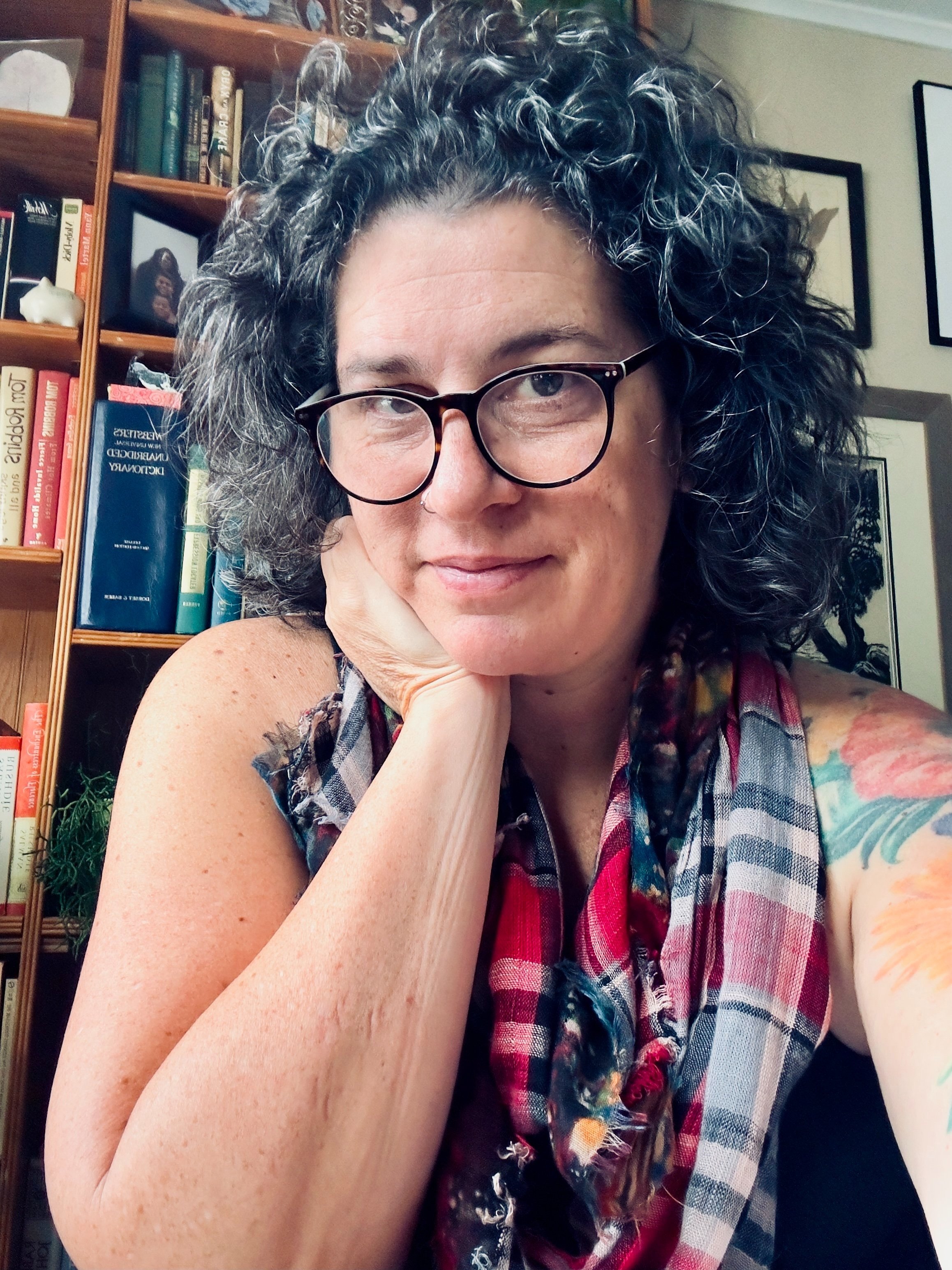 A woman with curly gray hair, glasses, and a tattoo on her upper arm, resting her chin on her hand, sitting in front of a bookshelf filled with books and framed photos.