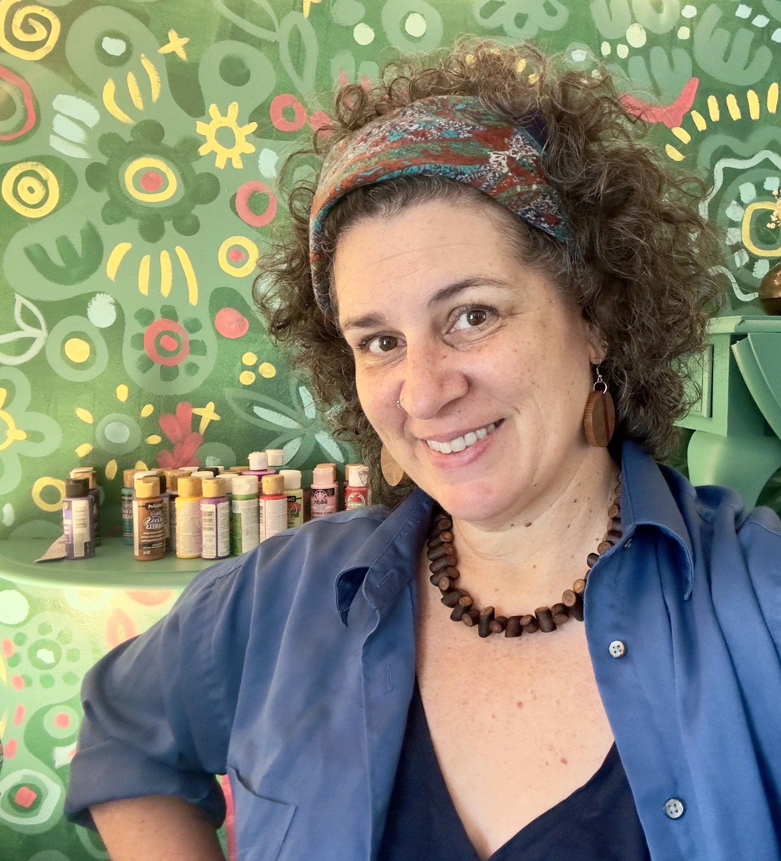 A woman with curly hair wearing a colorful headband, brown wooden earrings, and a beaded necklace, smiling in front of a green floral painted background and a shelf with vitamin bottles.