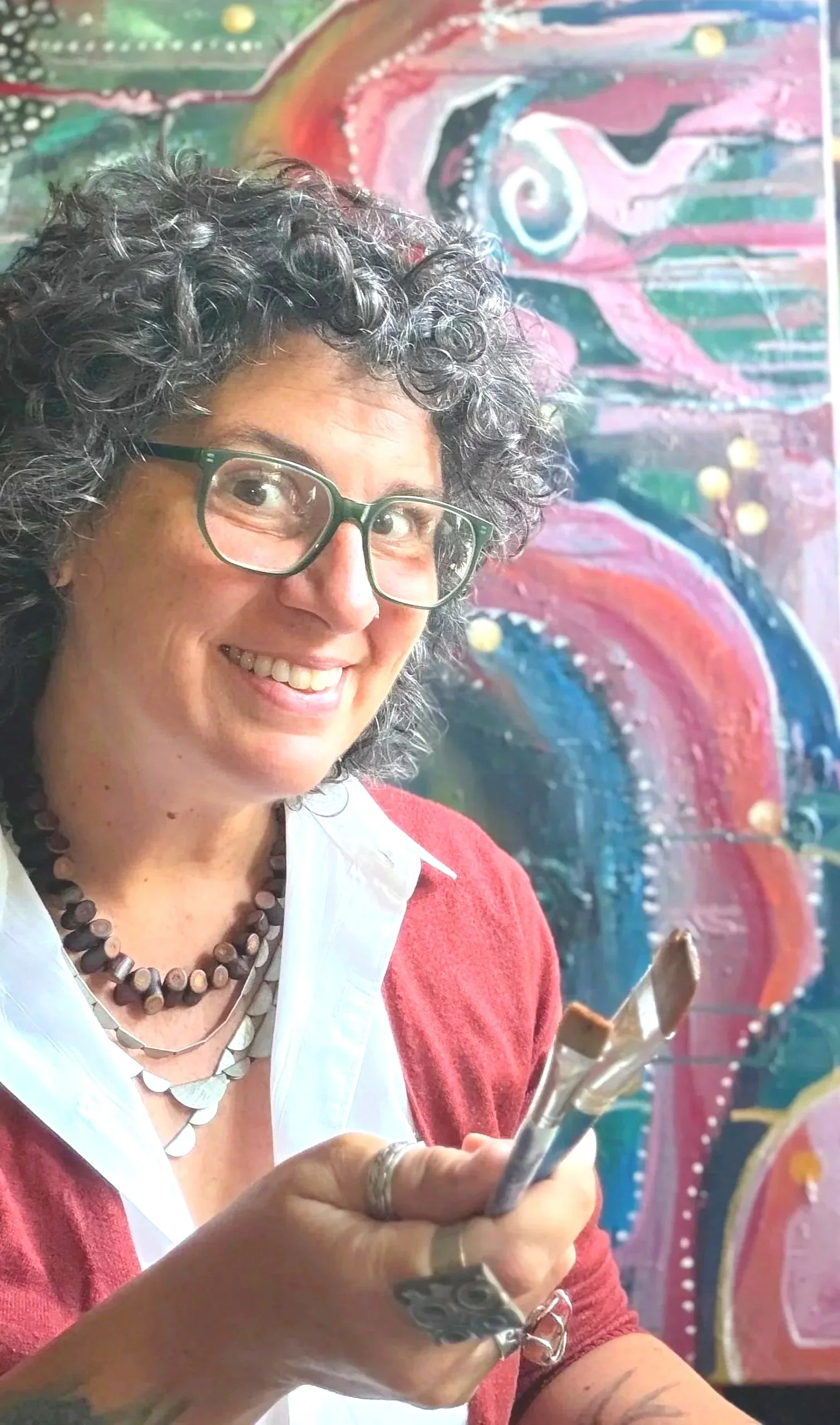 A woman with curly hair and glasses smiling while holding a paintbrush in front of a colorful abstract painting.