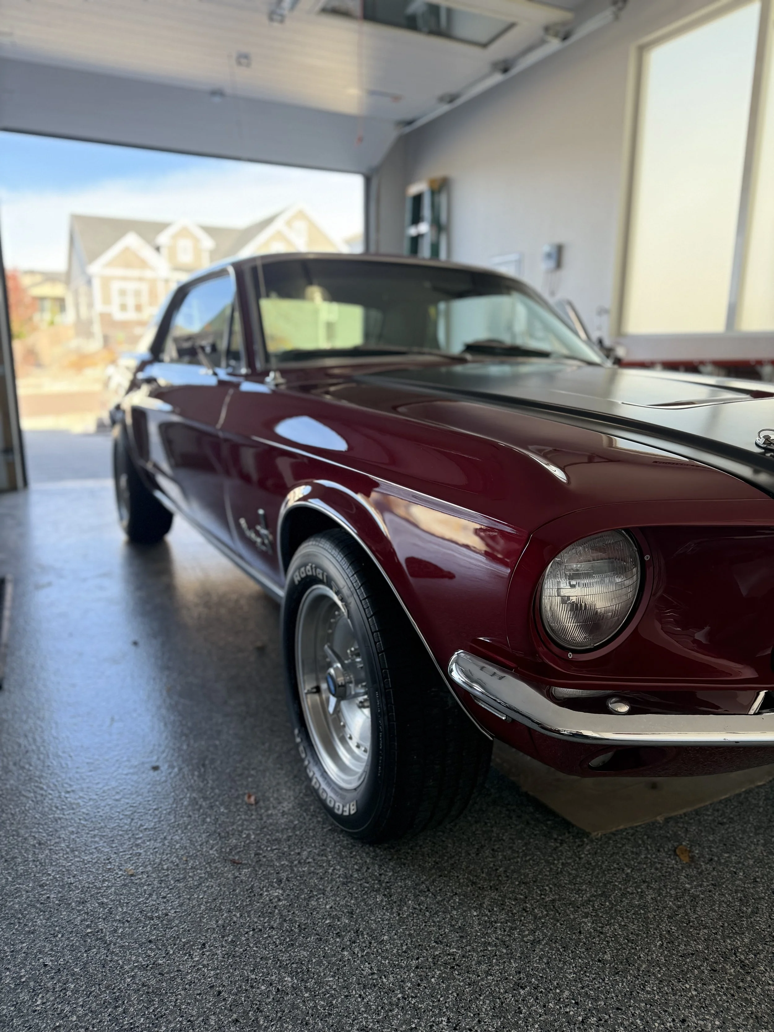 A red vintage sports car with black racing stripes parked inside a garage, with houses and a blue sky visible outside.