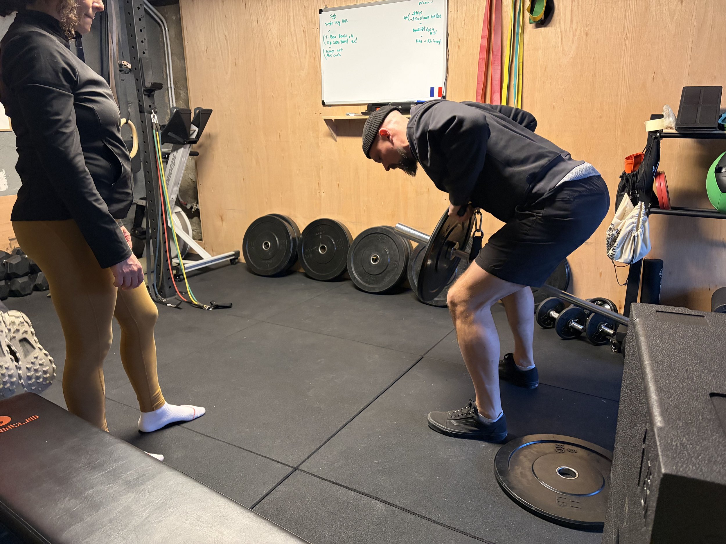 Un homme soulève un poids lors d'une séance d'entraînement dans une salle de sport, avec une femme à côté le regardant. La salle contient des barres, des poids, des bandes de résistance et autres équipements de musculation.