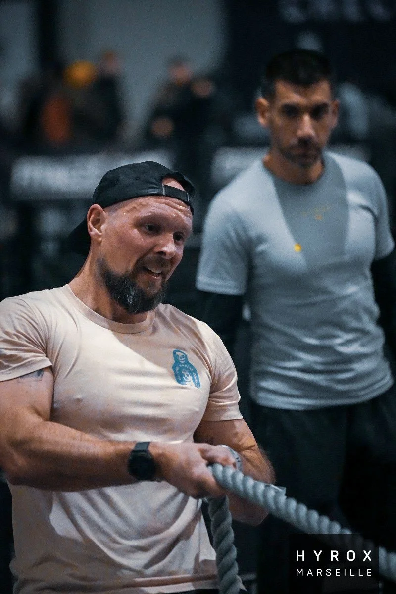 Deux hommes s'entraînent avec une corde dans une salle de sport, en arrière-plan d'autres personnes s'exercent. Logo 'HYROX MARSEILLE' en bas à droite.
