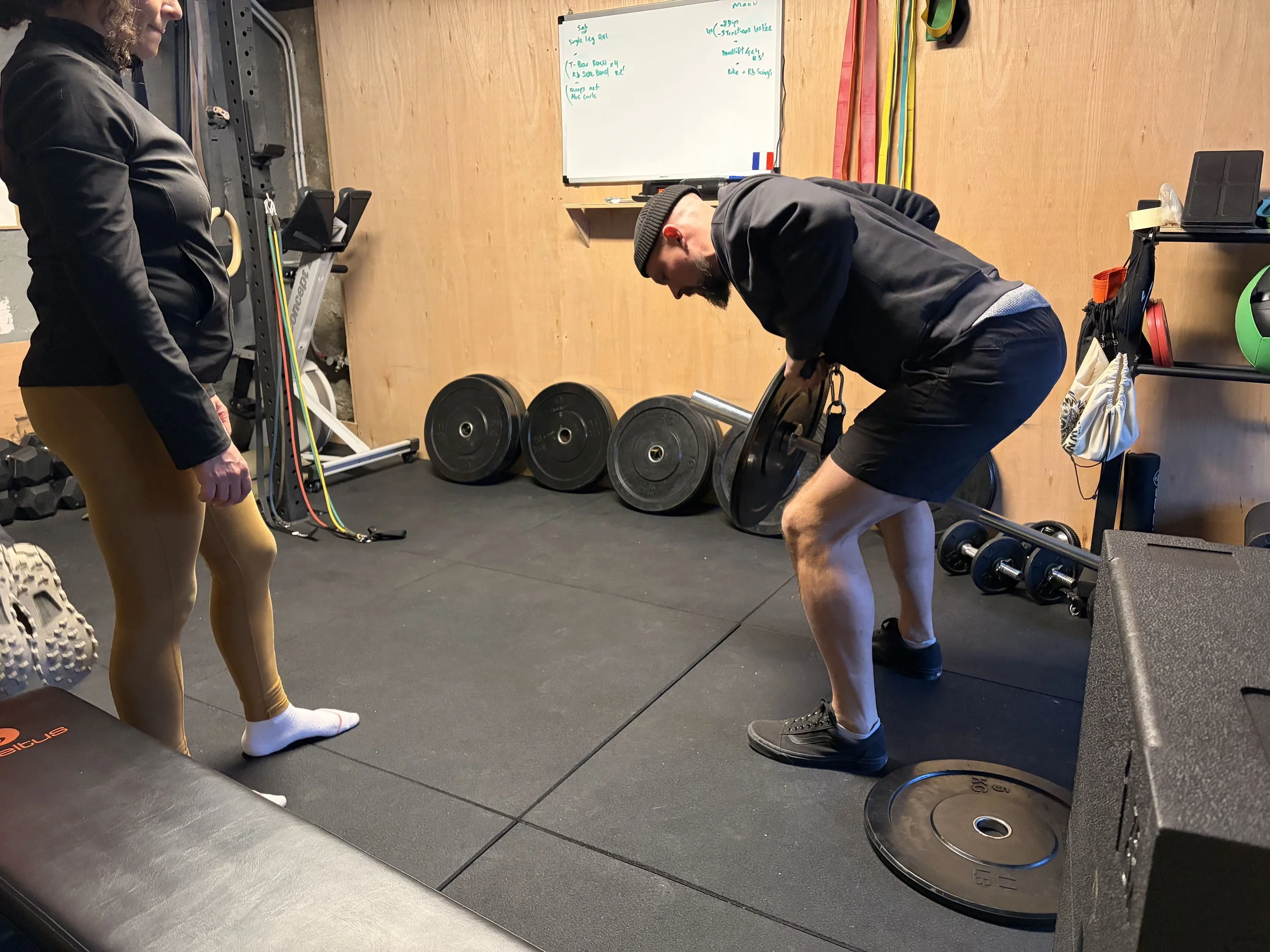 Deux personnes dans une salle de sport, une homme en train de soulever un disque de poids avec l'aide d'une femme qui l'observe. Il porte un bonnet, un sweat noir, des shorts noirs et des chaussures noires. La femme porte un léggings beige, un haut noir et des chaussettes blanches. L'arrière-plan montre des poids, des bandes de résistance, et un tableau blanc.