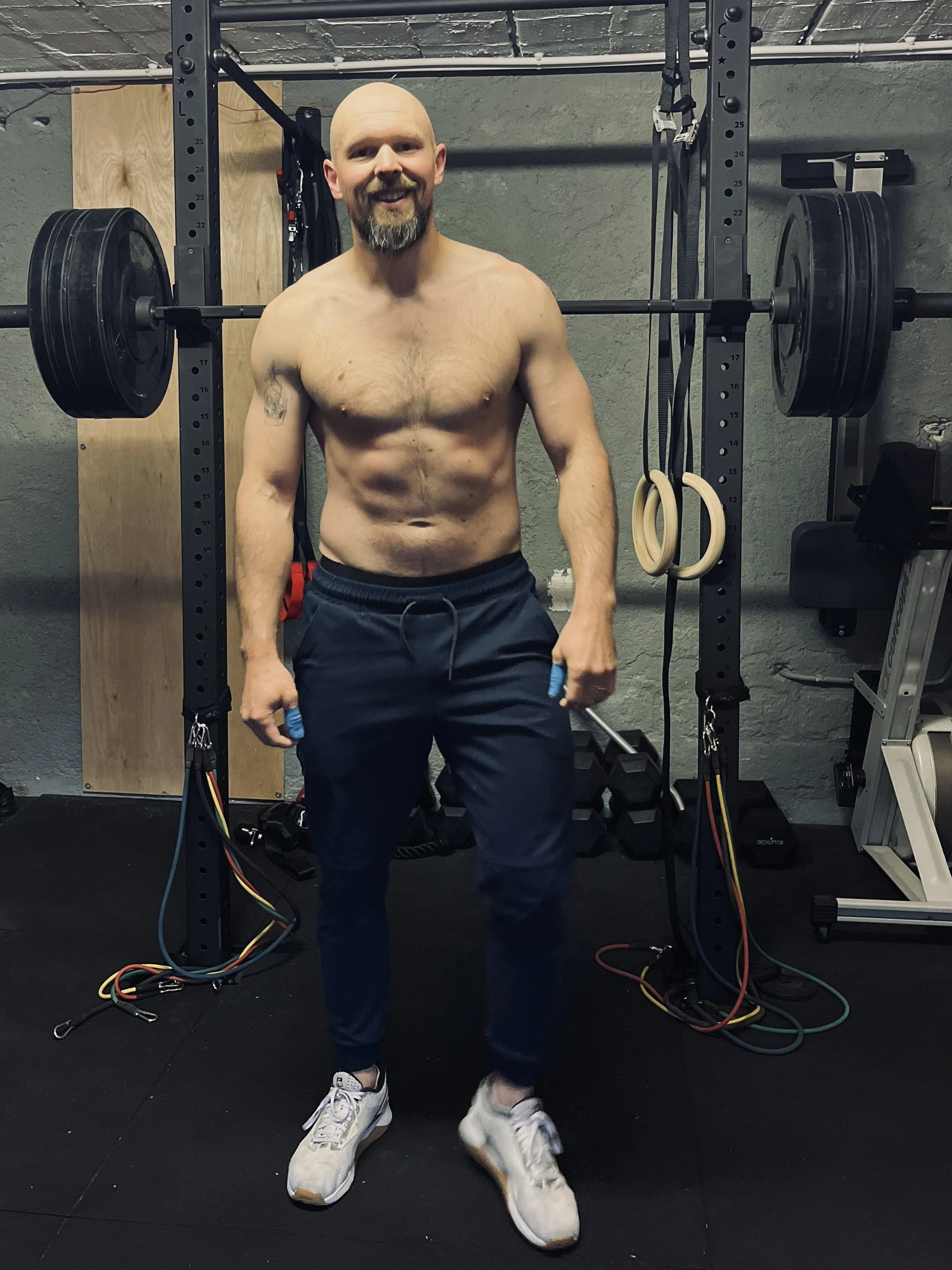 Homme musclé, sans chemise, portant un pantalon de sport blanc, debout dans une salle de sport devant un rack à poids avec des poids, des bandes de résistance, et des anneaux de gymnastique suspendus.