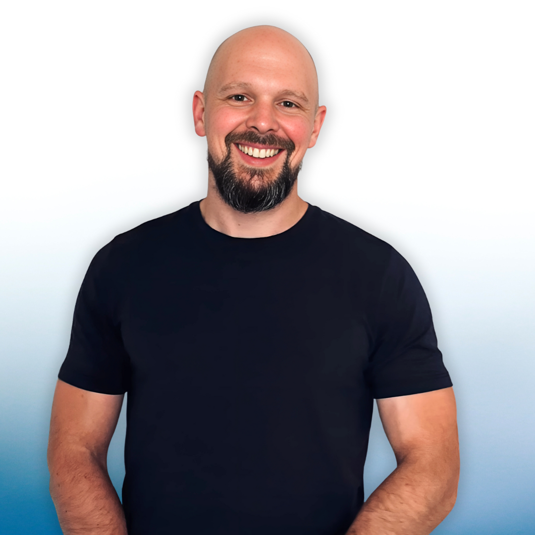 Un homme souriant avec une barbe et une tête rasée, portant un t-shirt noir, debout devant un fond blanc