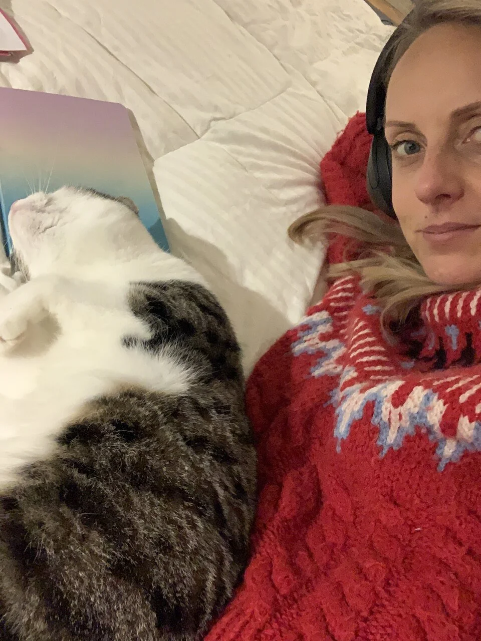 Woman in a cozy red sweater relaxing in bed with her cat, wearing headphones and enjoying a quiet moment at home.