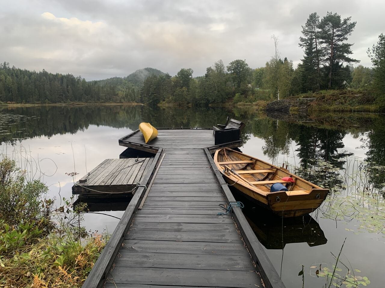 Empty wooden dock with a rowboat and chairs overlooking a calm lake surrounded by forest on an overcast autumn morning.