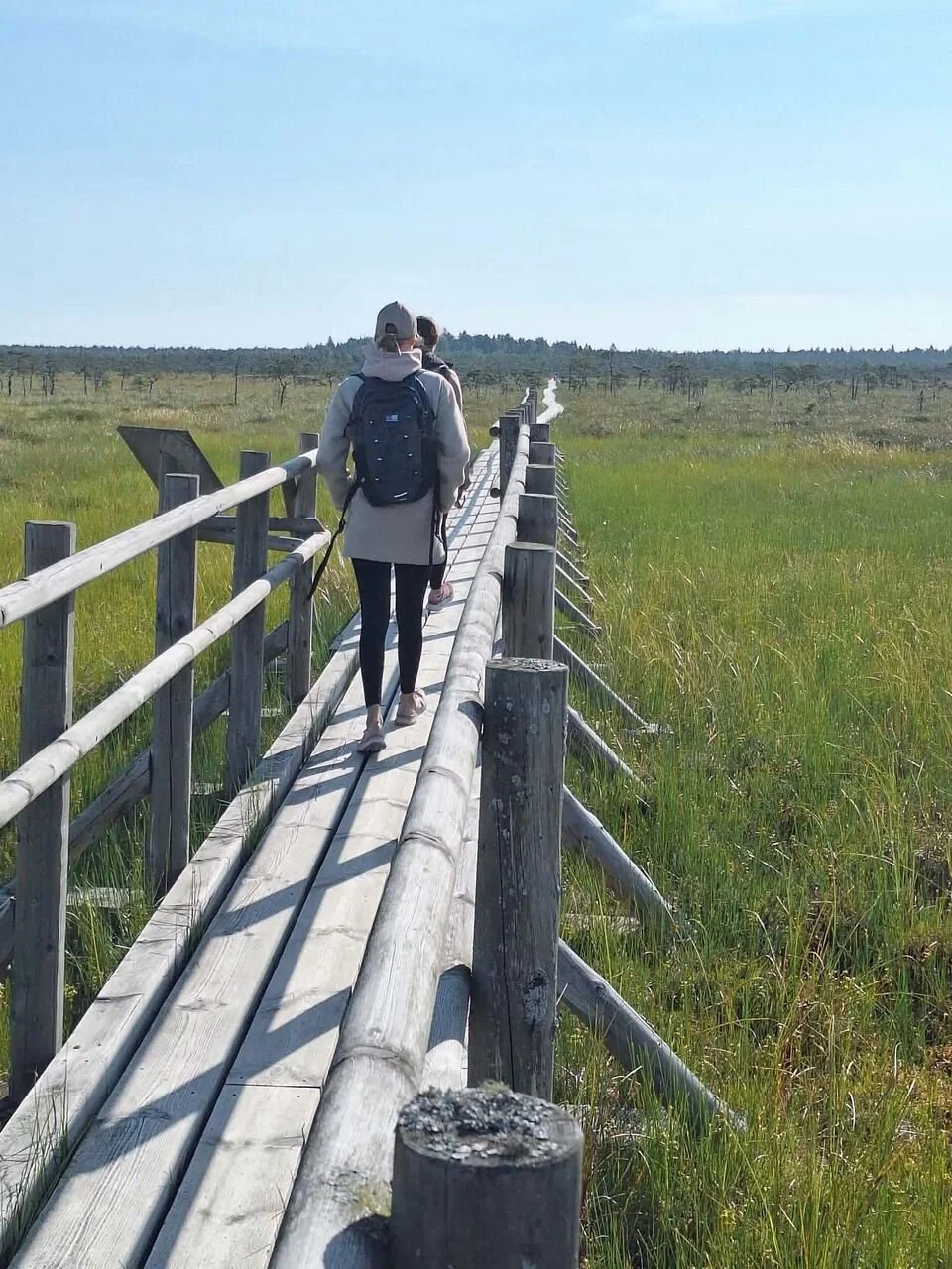 Two people walking along a wooden boardwalk through a wide, open bog landscape.