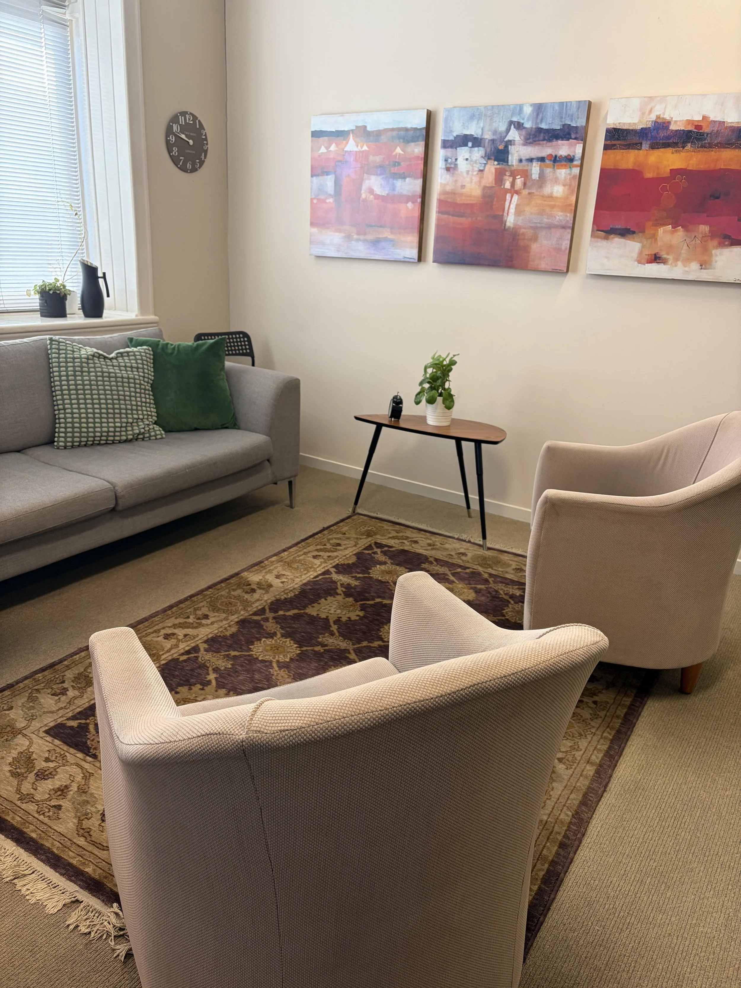 A cozy living room with beige and gray furniture, a patterned rug, art paintings on the wall, and decorative pillows.