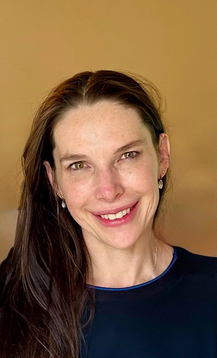 Close-up photo of a smiling woman with long brown hair, wearing earrings, and a dark top with blue trim, against a beige background.