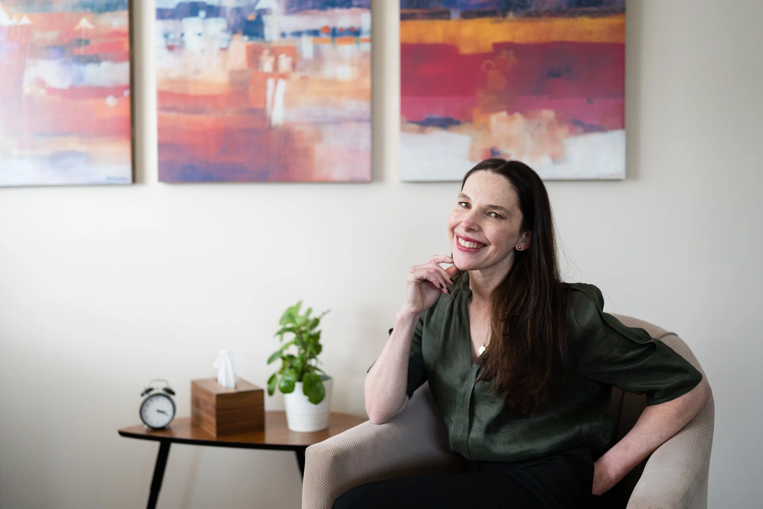 A woman with dark hair and a green blouse sitting on a beige chair, smiling and resting her chin on her hand, in a room with abstract colorful paintings on the wall, a wooden side table with a potted plant, a clock, and a tissue box.