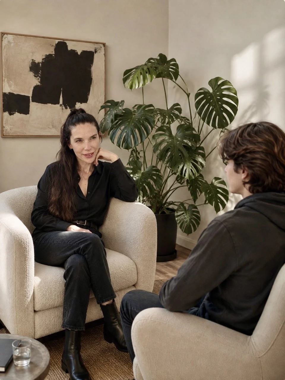 A woman and a man having a conversation in a cozy room, with a large potted plant and an abstract painting on the wall behind them.