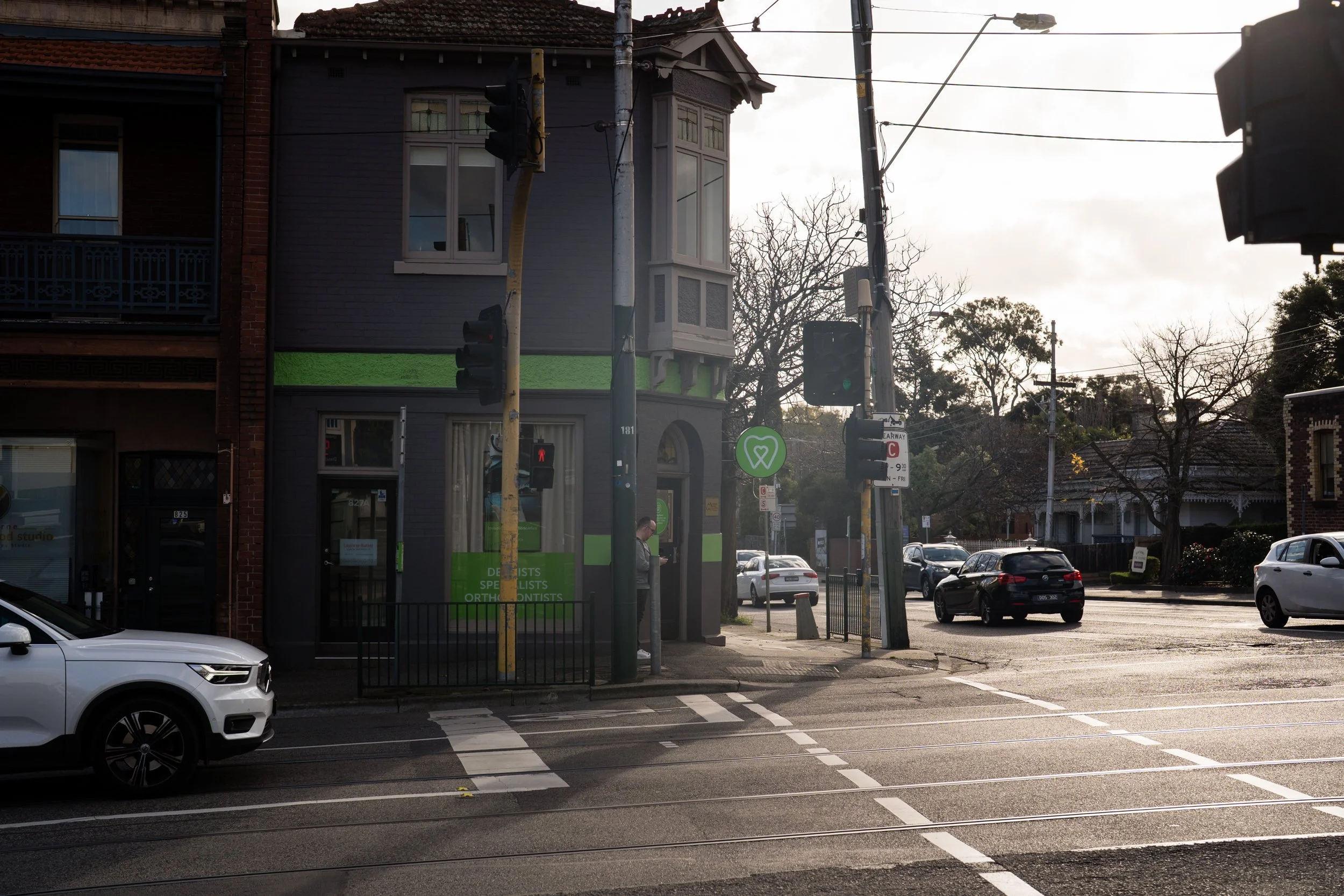 Street intersection with a gray building featuring large windows and a green sign, traffic lights, several parked cars, and a person crossing the street.