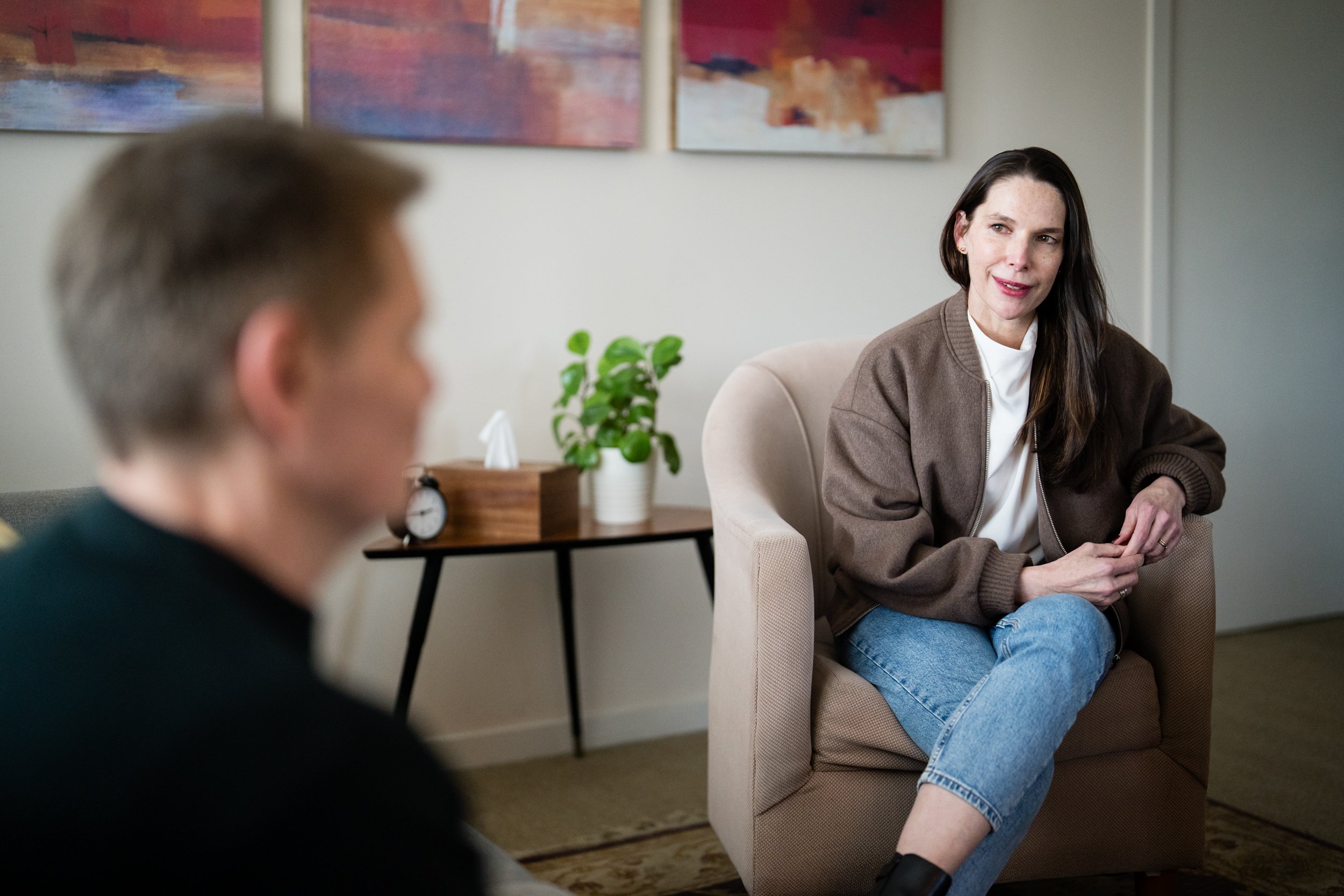 A woman with long dark hair speaking, sitting on a beige armchair, with a man blurred in the foreground, in a room with abstract art on the wall and a small table with a plant, clock, and tissue box.