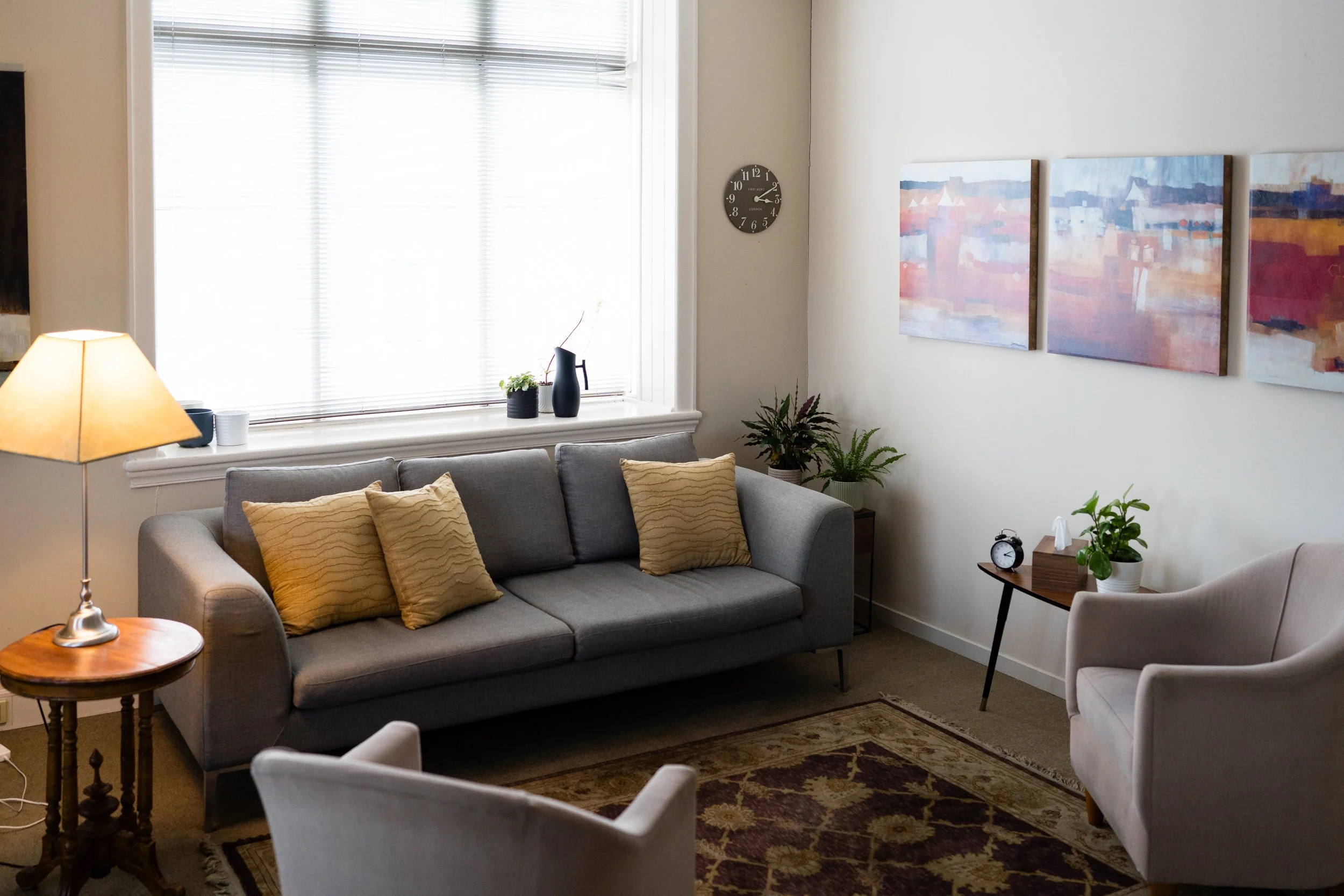 Living room with gray couch, beige pillows, wooden side table, white armchair, potted plants, abstract paintings on wall, window with blinds, and decorative vases on window sill.
