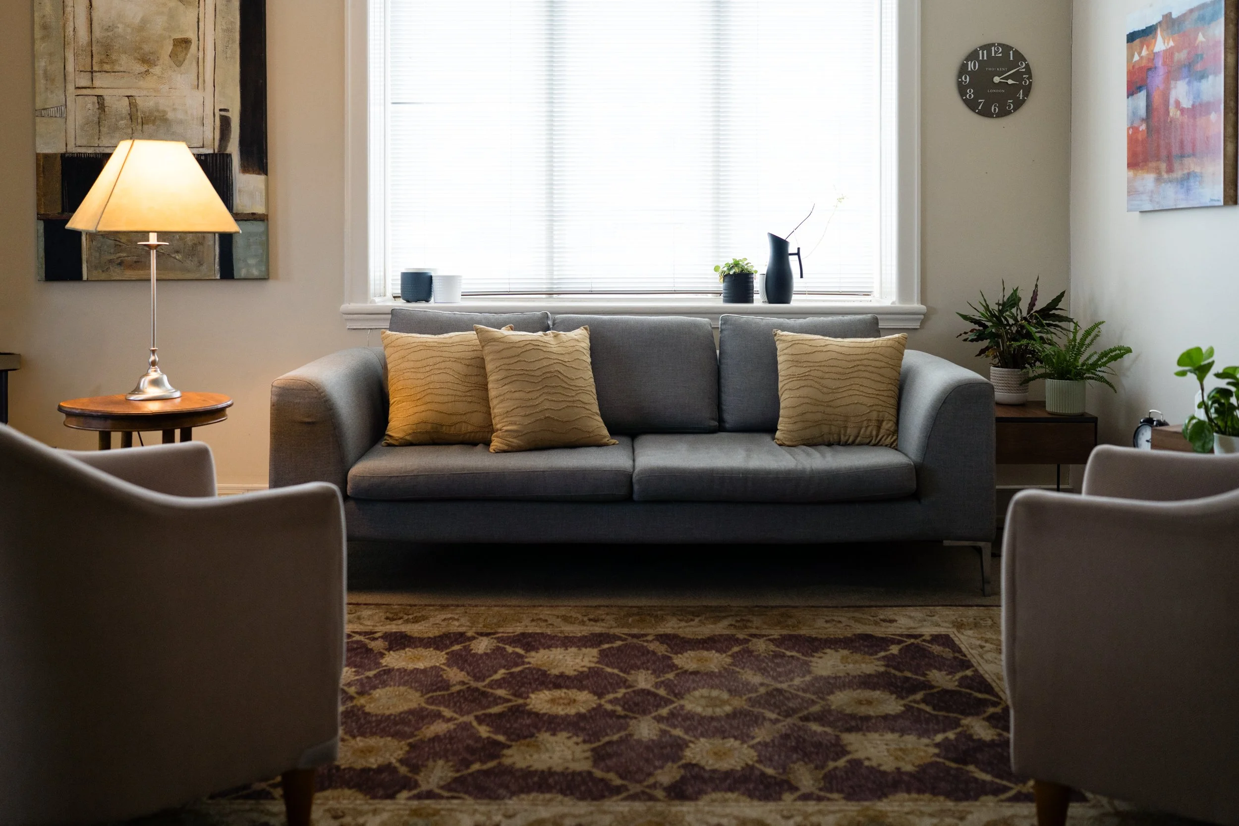 Living room with a gray sofa, yellow pillows, a wooden side table with a lamp, a patterned rug, a window with blinds, and various decorative items including plants and artwork.