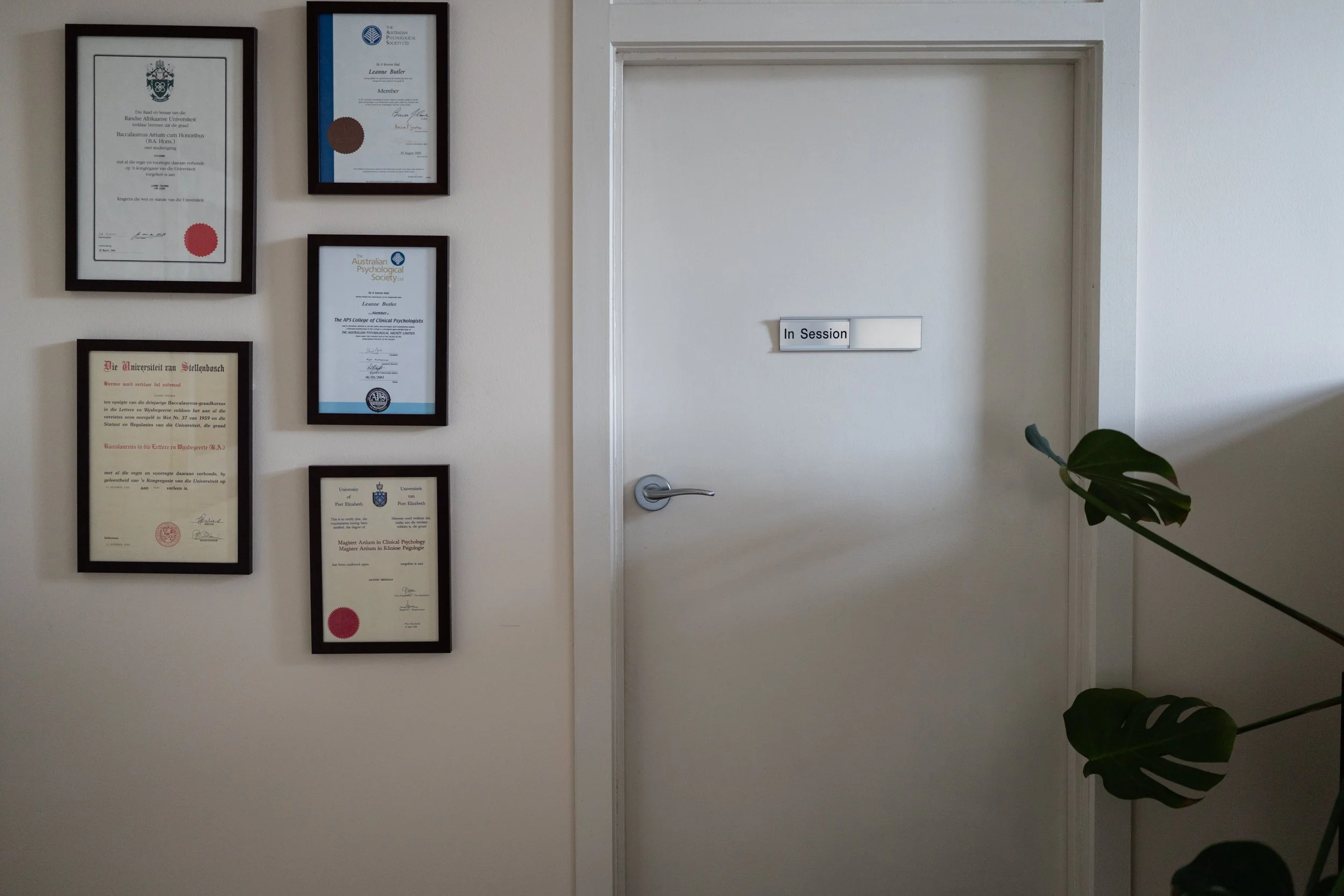 A white door with a small white sign that reads 'In Session' in black text. To the left of the door, there are various framed certificates and diplomas mounted on the wall. A green plant is partially visible in the bottom right corner.