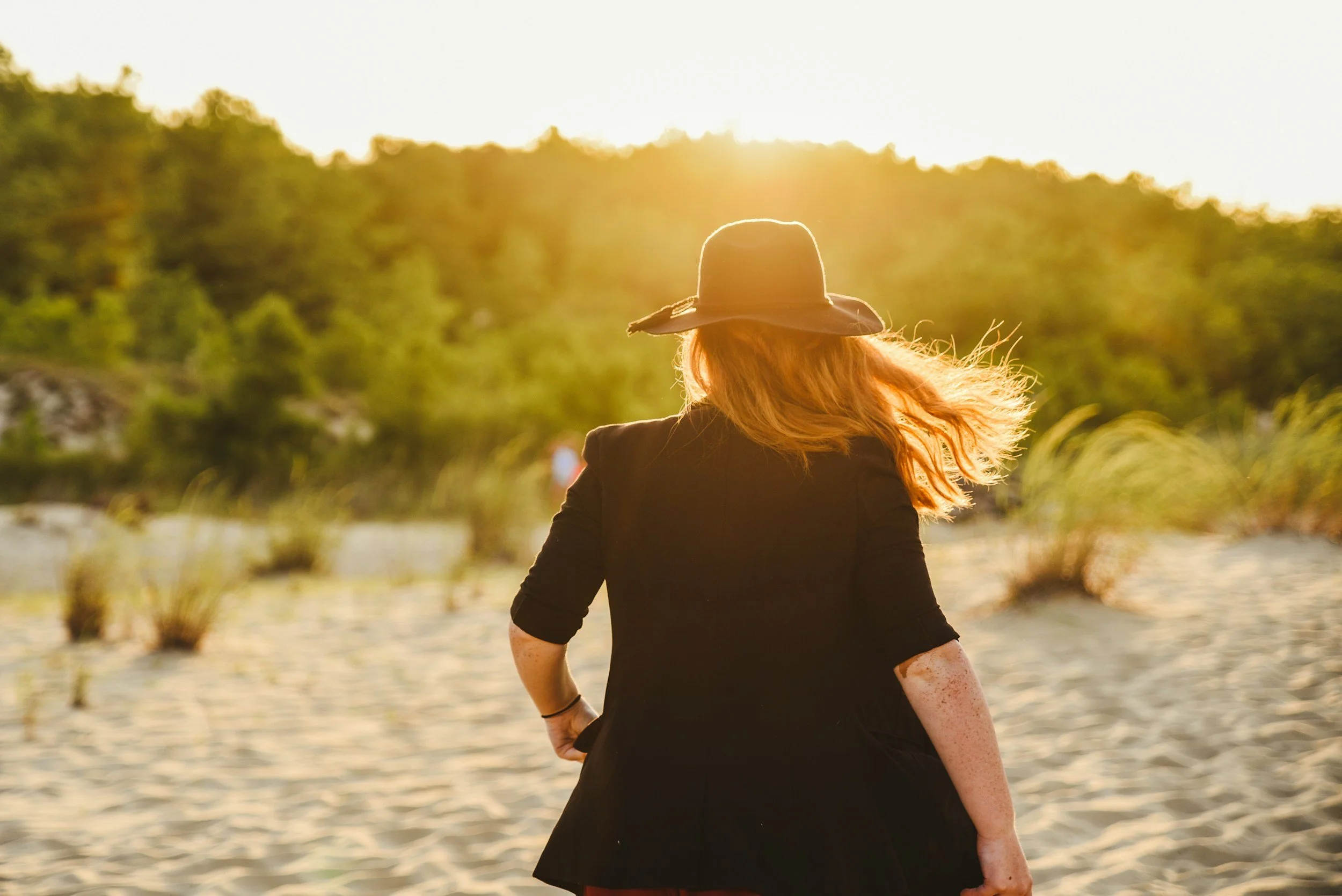 Woman with red hair wearing a black jacket and a wide-brimmed hat walking on a sandy beach during sunset