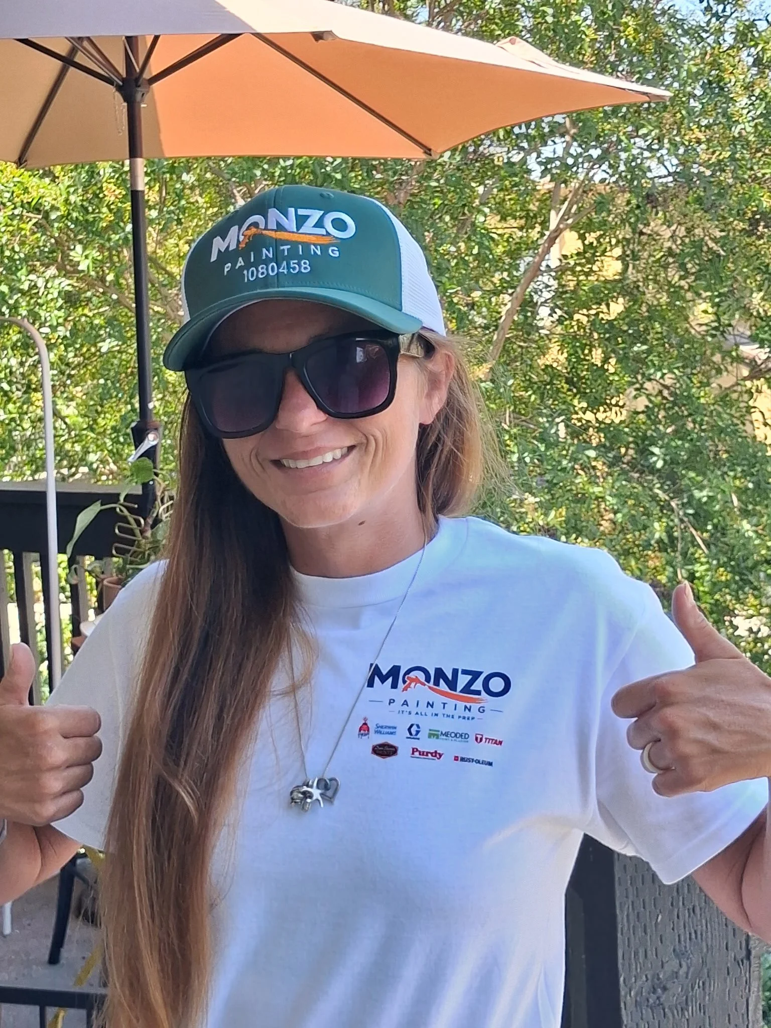 Woman smiling, wearing sunglasses and a blue cap with 'MONZO PAINTING' logo, giving a thumbs-up, standing outdoors under a patio umbrella surrounded by trees.